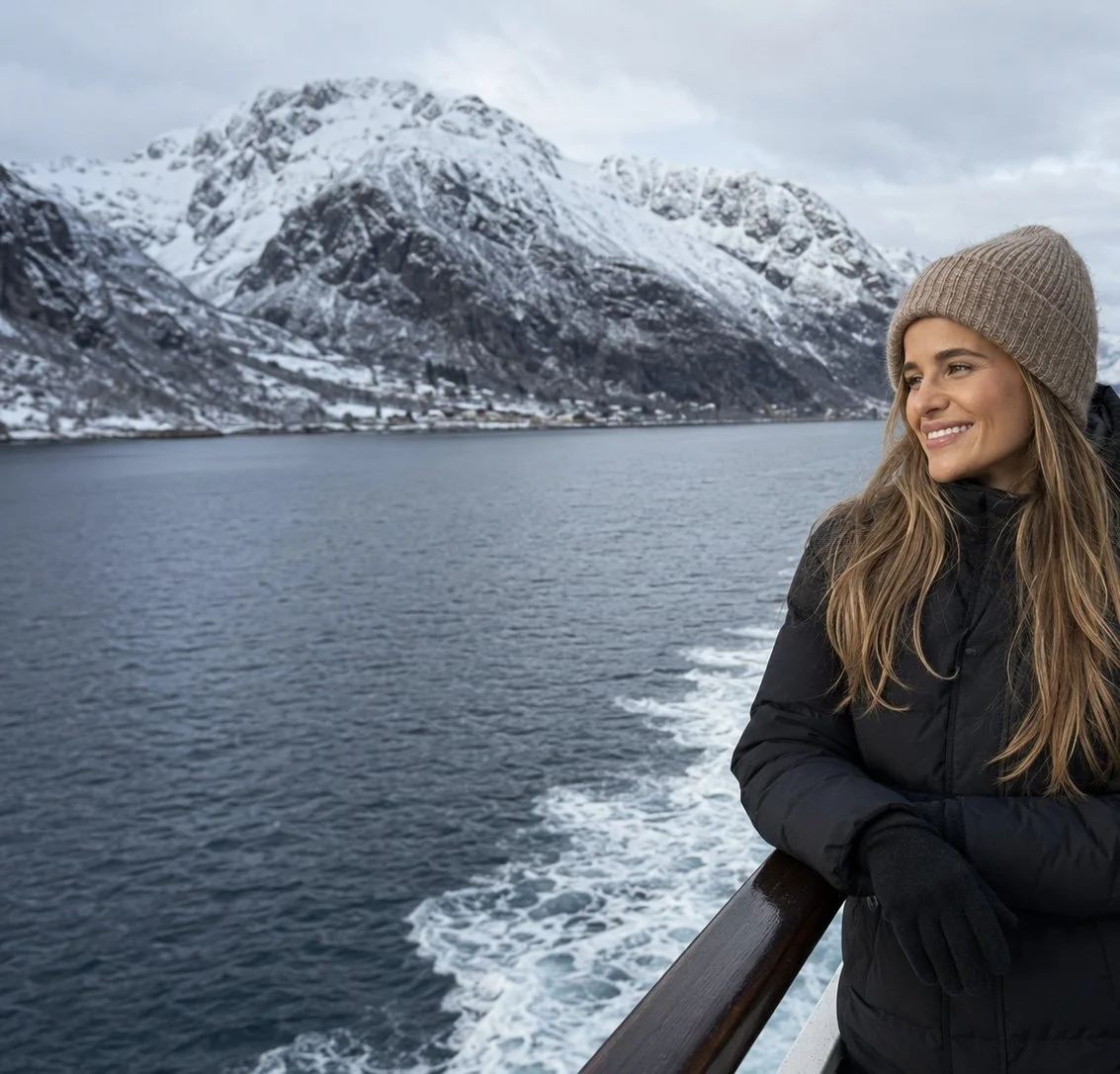 A woman standing on deck smiling, looking at the snowy Norwegian mountians, wearing a thick winter parka and a wool hat.