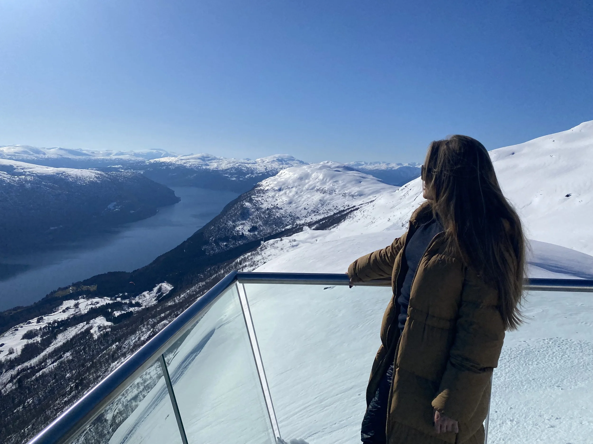 Woman standing at the top of Loen Skylift in April sunshine, with snow-covered mountains behind her and a snow-free valley below.