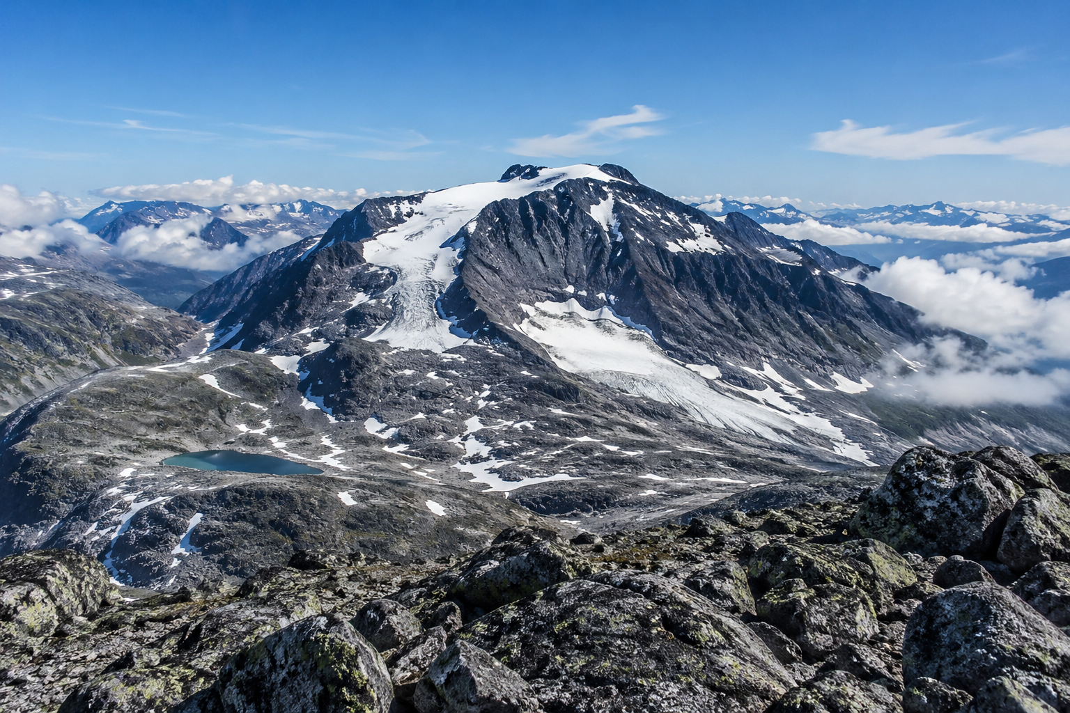 A picture from the top of Galdhøpiggen, with snow covered tops and blue skies