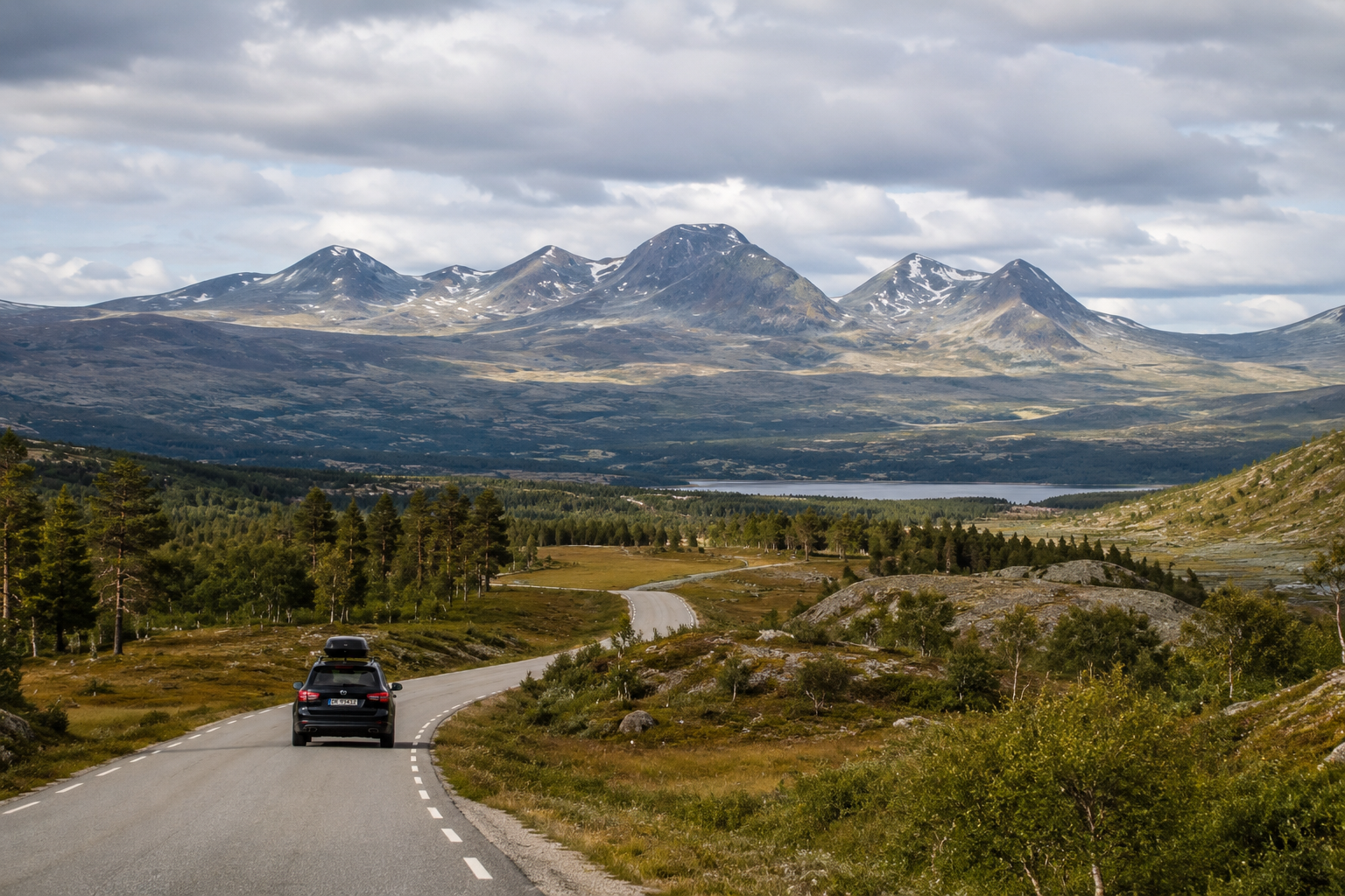 Scenic mountain road through Rondane, Norway, with a car driving towards the open landscape, forest, lake and rounded mountain peaks beneath a cloudy summer sky.