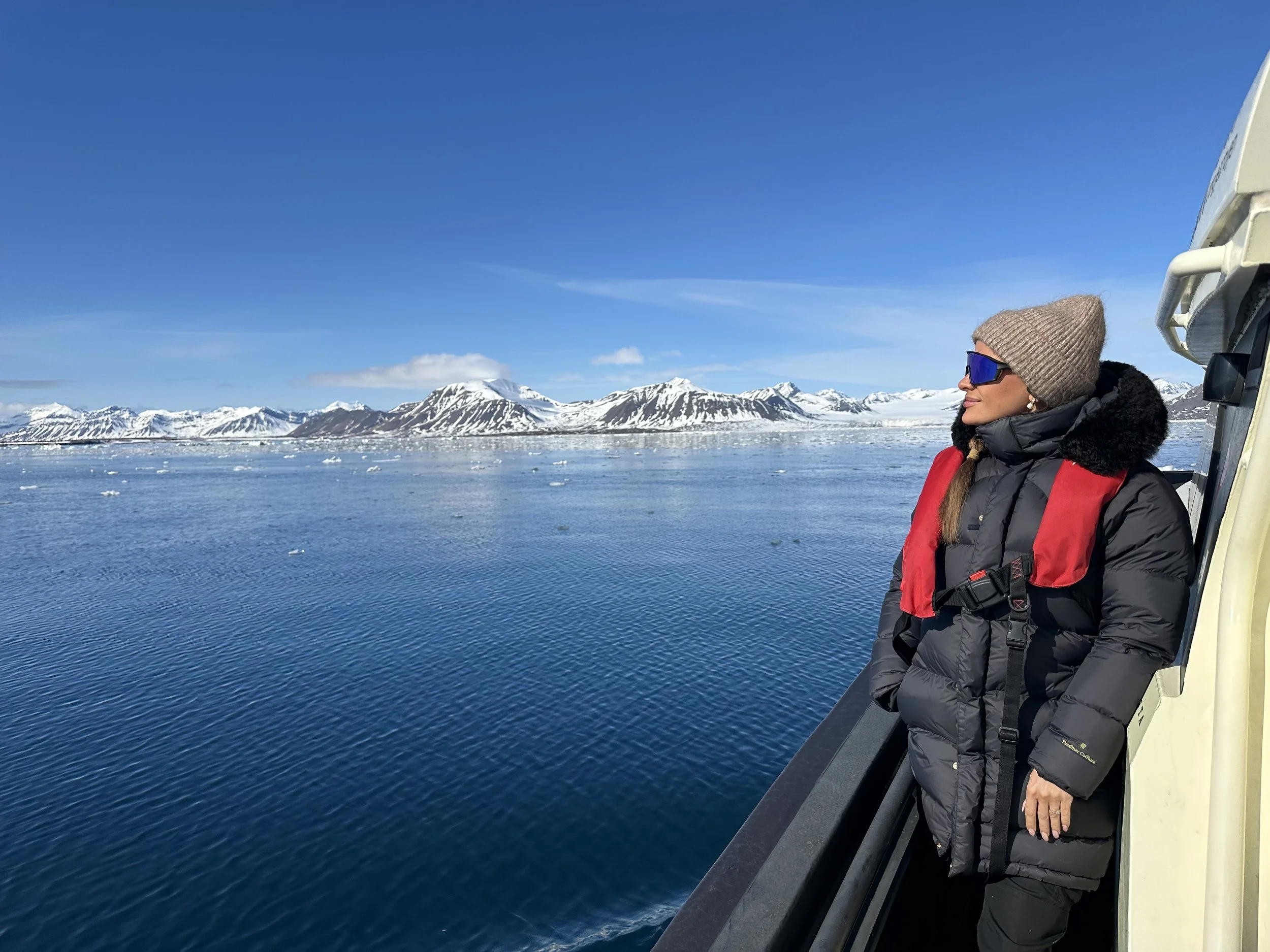 A woman standing on deck looking out on the snowy mountain tops wearing a wool hat, thick coat and sunglasses.