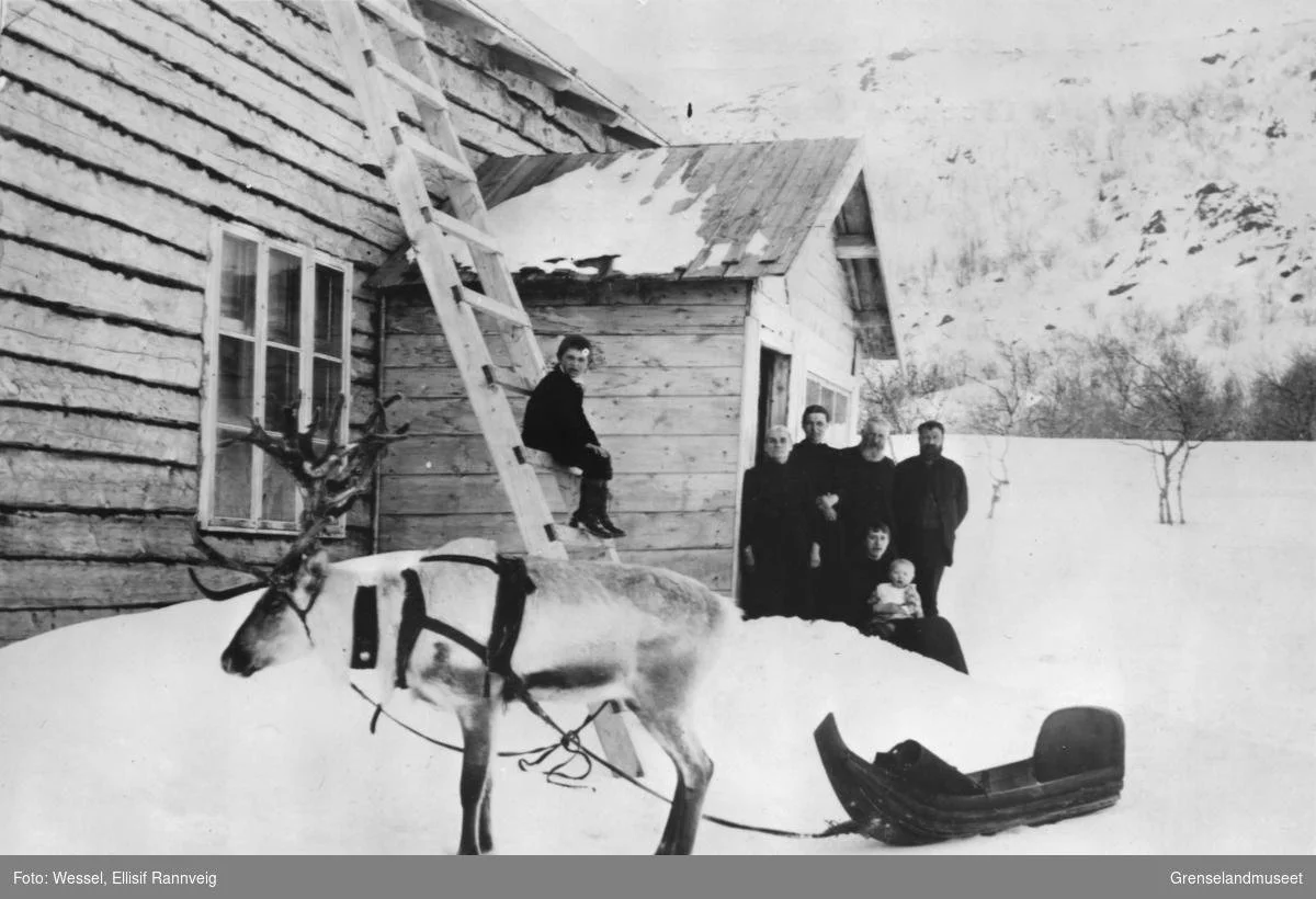 A black and white photo of a family standing outside their house on their homestead in Kirkenes, with a reindeer with a little sled.
