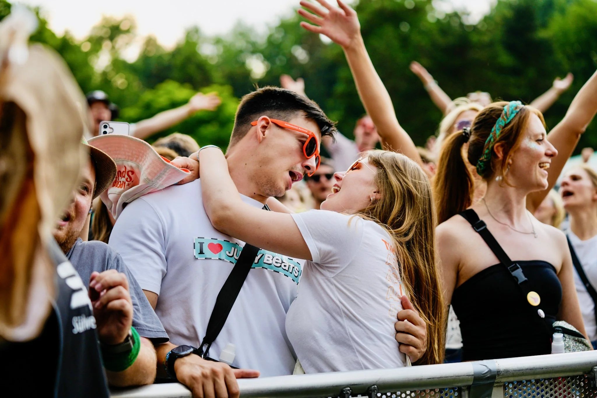 A crowd of people at an outdoor event, with a young man and woman in the foreground embracing each other and smiling. Many have their arms raised in celebration, and there are trees in the background.