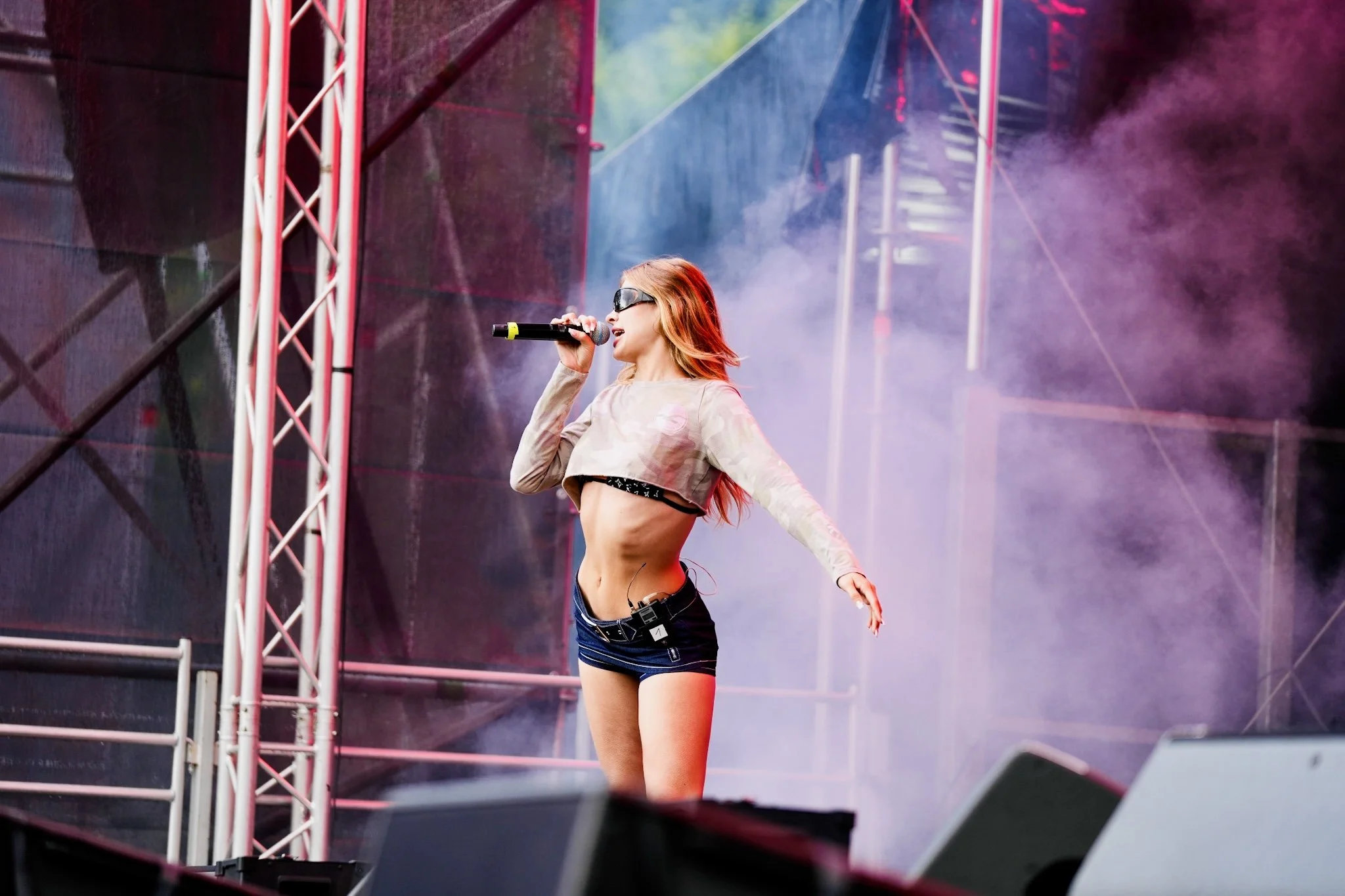 A female performer with red hair singing into a microphone on stage during a concert, wearing sunglasses, a cropped top, and shorts, with stage equipment and colorful lights in the background.