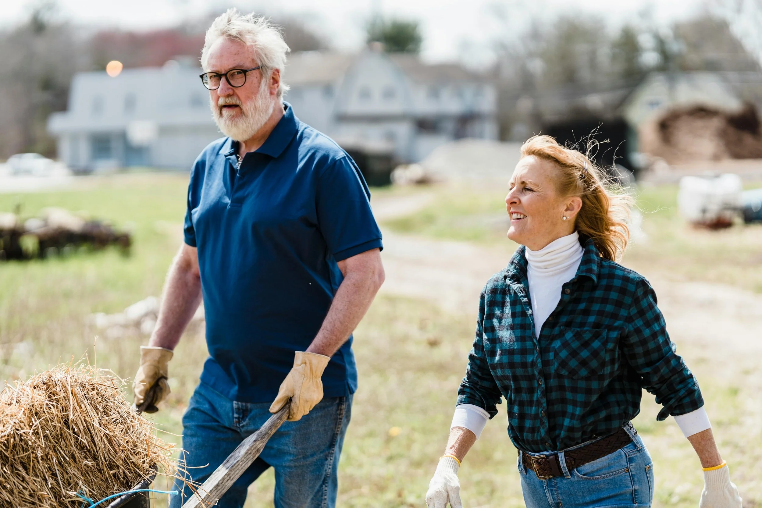A mature man with gray hair, glasses, and a beard wearing a deep blue shirt pushes a wheel barrel while his wife dressed in a white turtle neck and green and black buffalo plaid shirt walks along him smiling