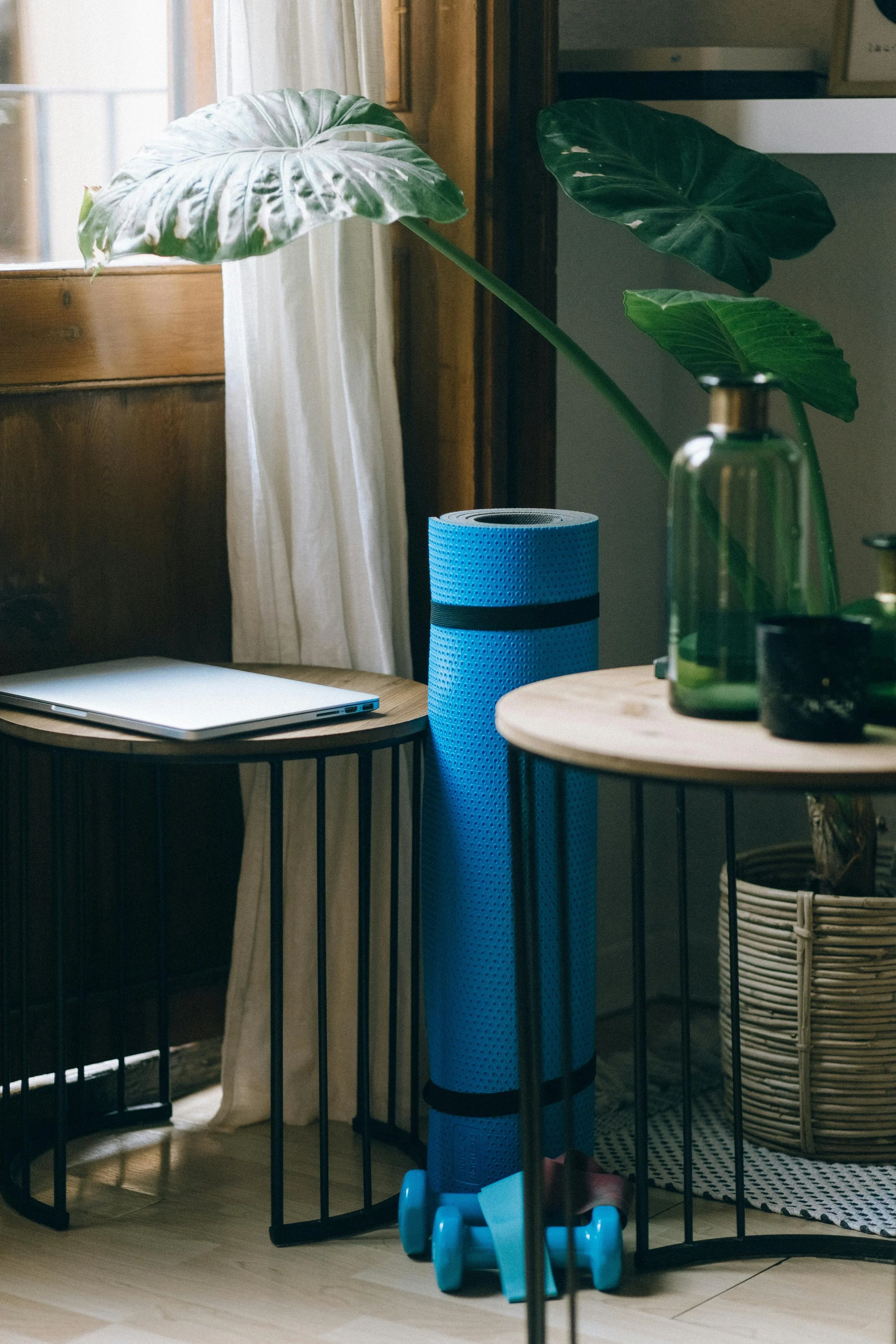 Two metal side tables with wooden tops sit next to each other with a royal blue yoga mat between with a laptop on the left table and a vase on the other. Light filters in through a window while a plant stretches in the background to reach the light.