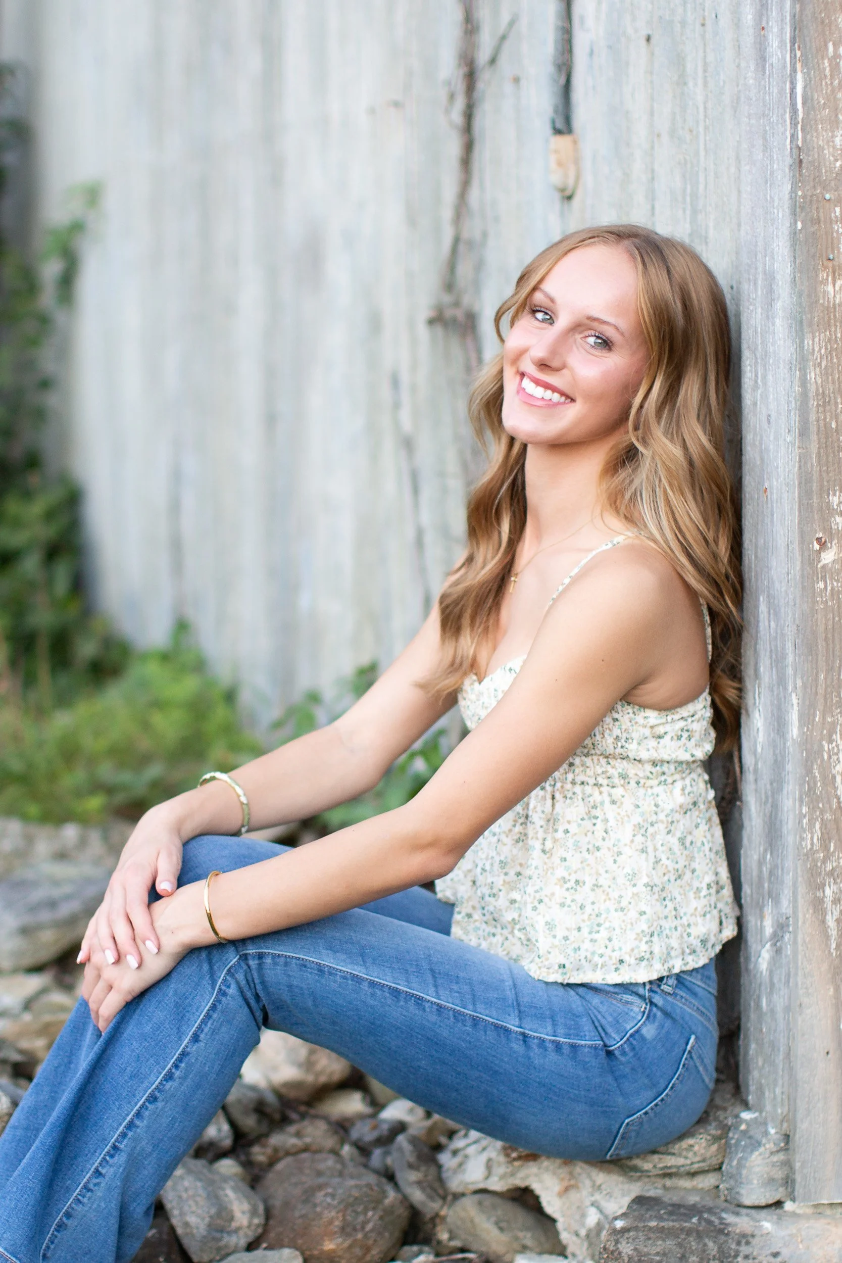 Casual senior portrait of Audrey wearing jeans and a floral top beside a weathered barn.
