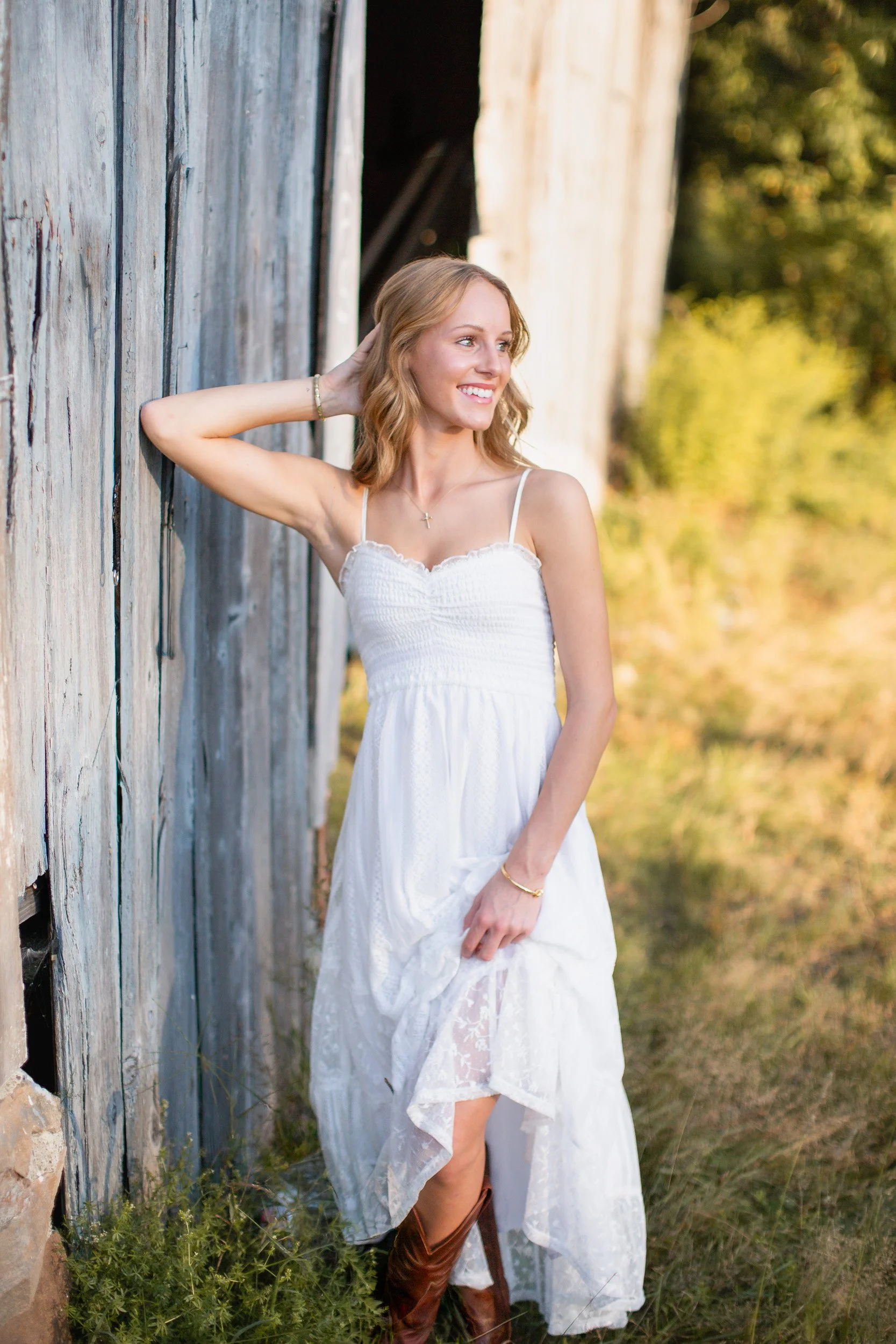 Outdoor senior portrait of Audrey in a white dress beside a weathered blue-gray barn.
