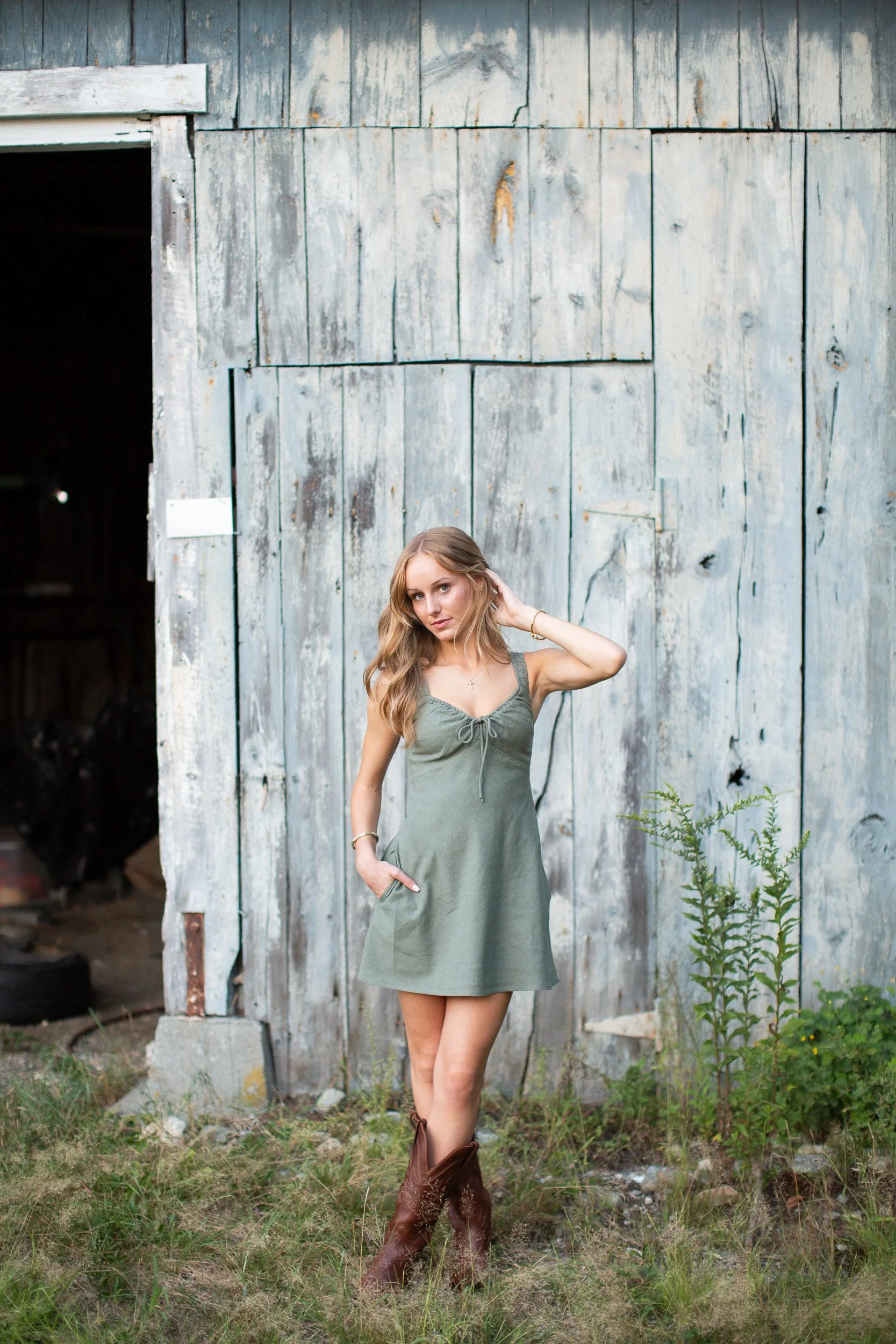 Senior girl in a green dress and cowboy boots posing in front of a rustic weathered barn.