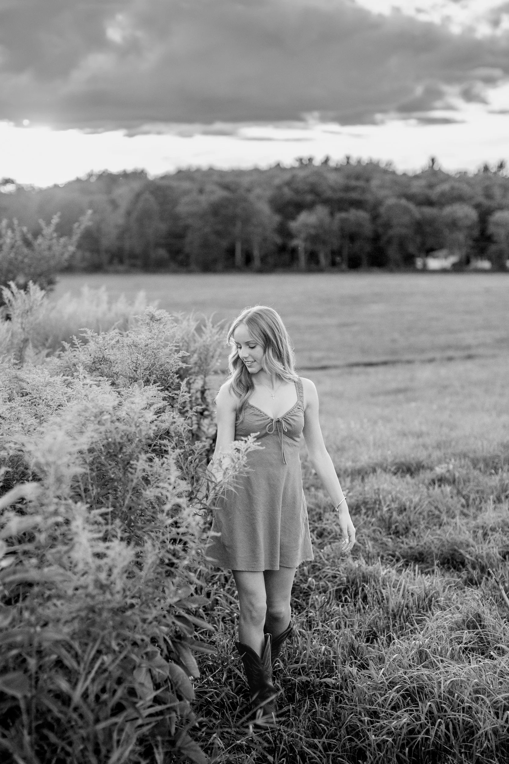 Black-and-white senior portrait of Audrey walking through tall grasses beside an open field.