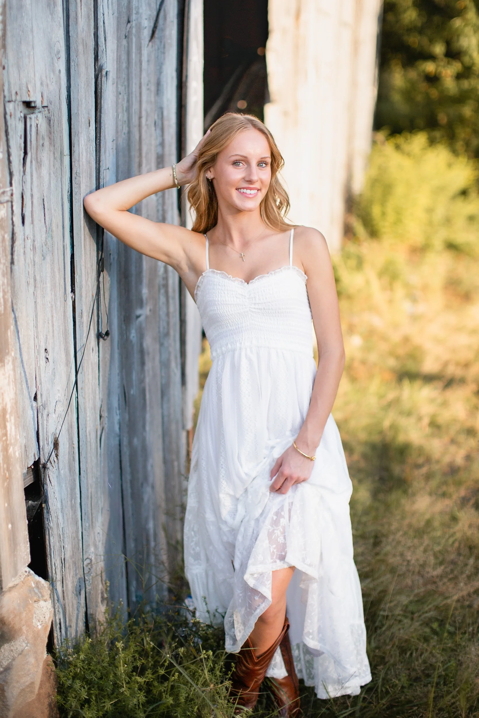 Audrey, a Mansfield senior, wearing a white dress and cowboy boots beside a weathered barn for her Massachusetts senior photos.