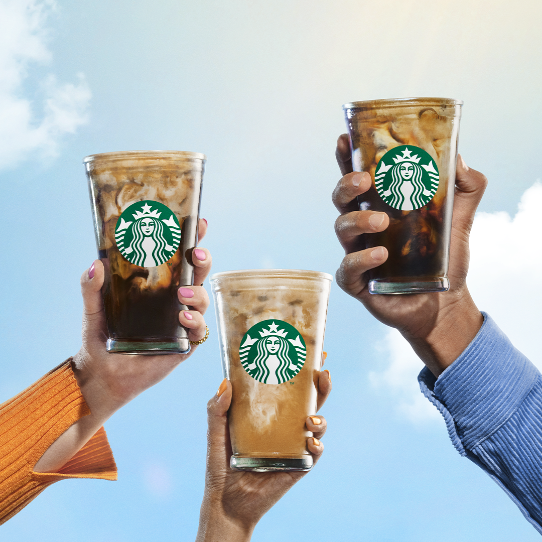 Three hands holding Starbucks iced coffee drinks with ice against a partly cloudy sky.