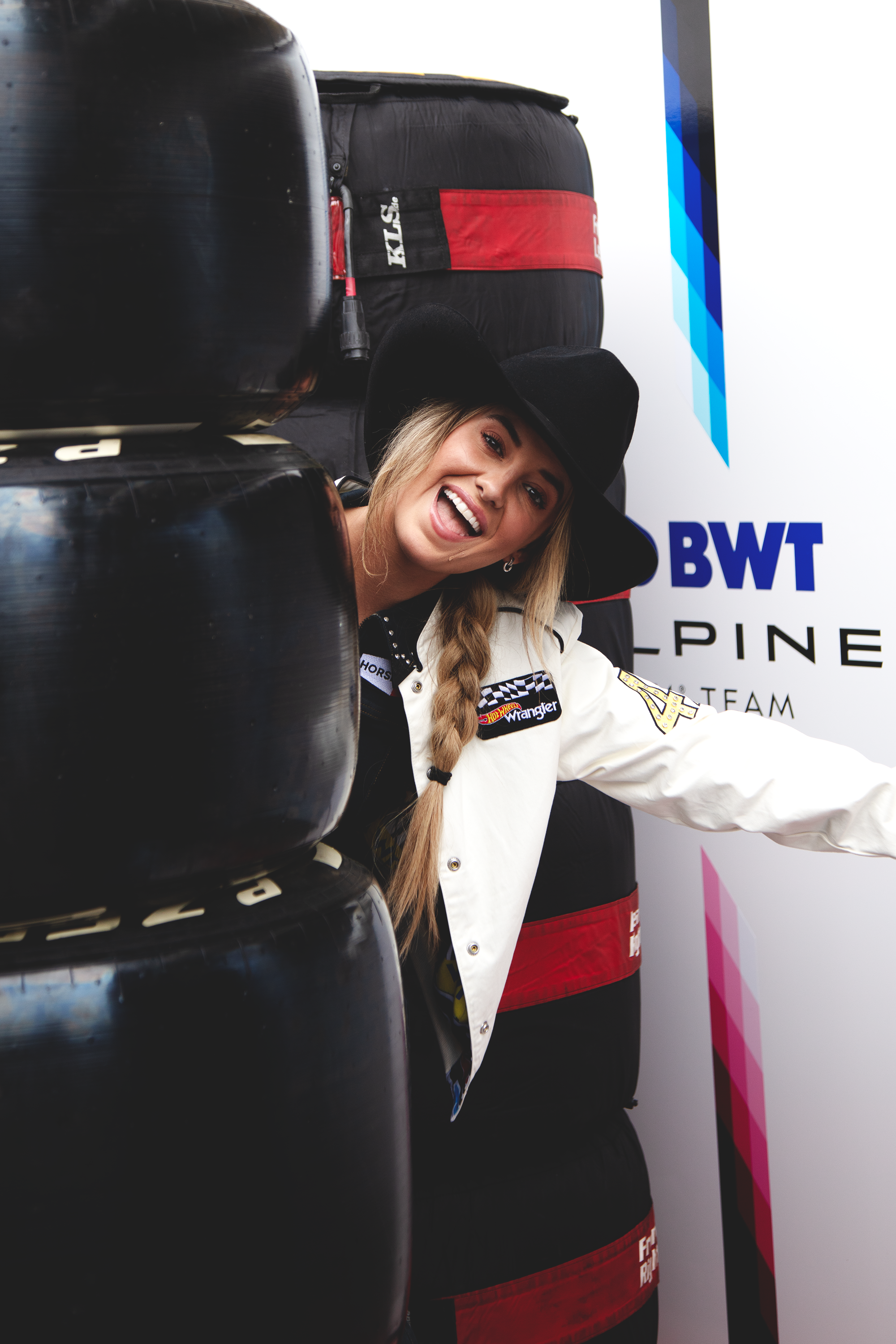 A smiling woman with a braid and wearing a black cowboy hat peeking out from behind a stack of tires in a racing environment, with a backdrop that includes a logo and a racing team name.