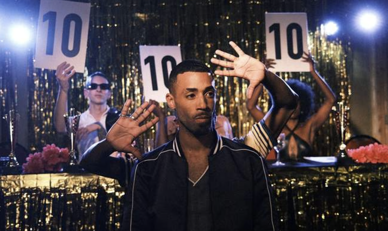 A group of people celebrating at a party with a glamorous background of shiny gold tiles, holding signs with a number 10, and a man in the foreground making a gesture with his hands.