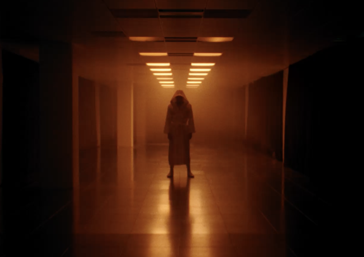 A person standing alone in a dimly lit corridor with rectangular ceiling lights casting a warm glow.