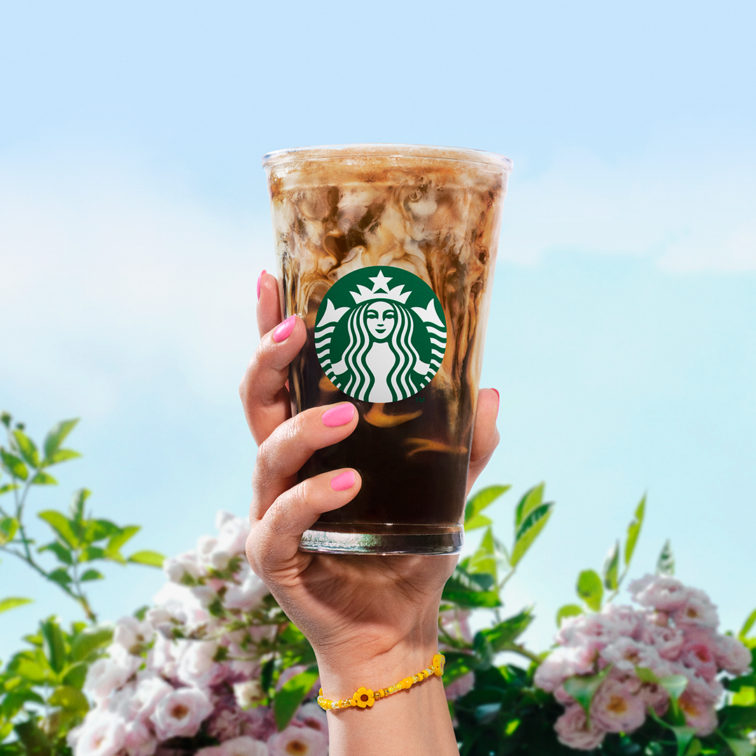 A hand holding a large iced coffee with the Starbucks logo, against a background of blue sky and pink flowers.