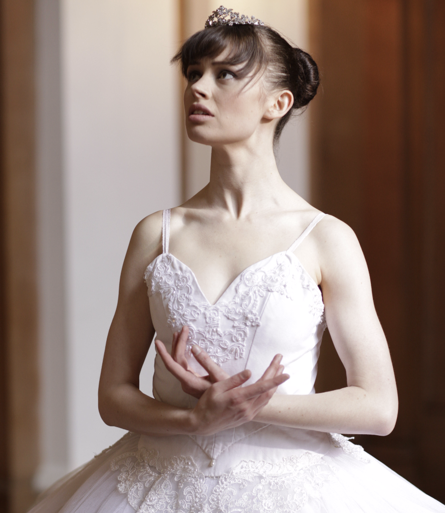 A young woman dressed in a white ballet outfit with lace details, wearing a tiara, and standing indoors.