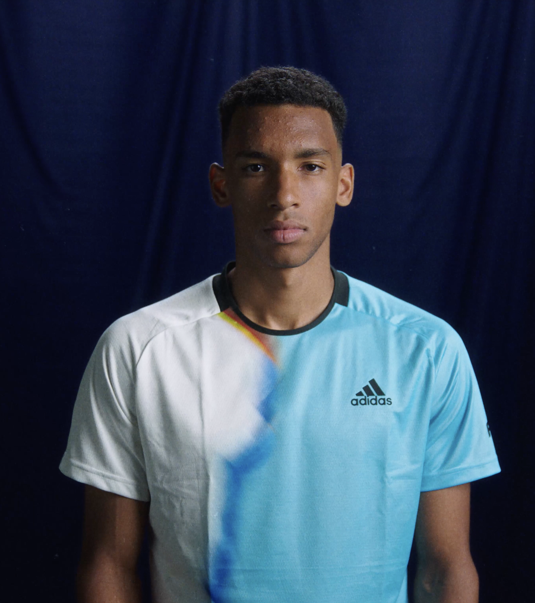 Felix  Auger Aliassime wearing a white and blue Adidas sports shirt standing against a dark blue background.