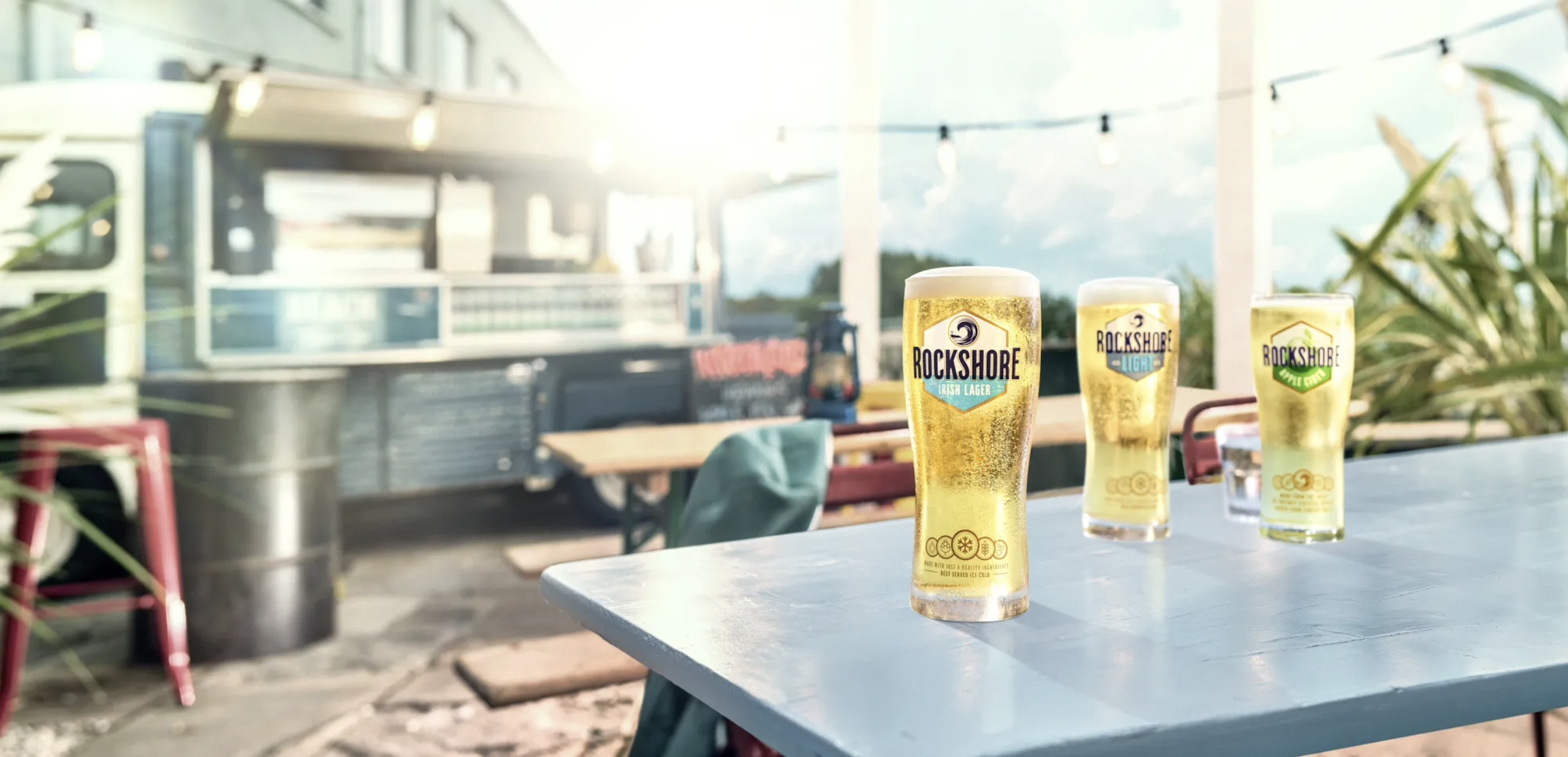 Three glasses of Rockshore Irish lager beer on an outdoor table with sunlight, a food truck in the background, and string lights overhead.