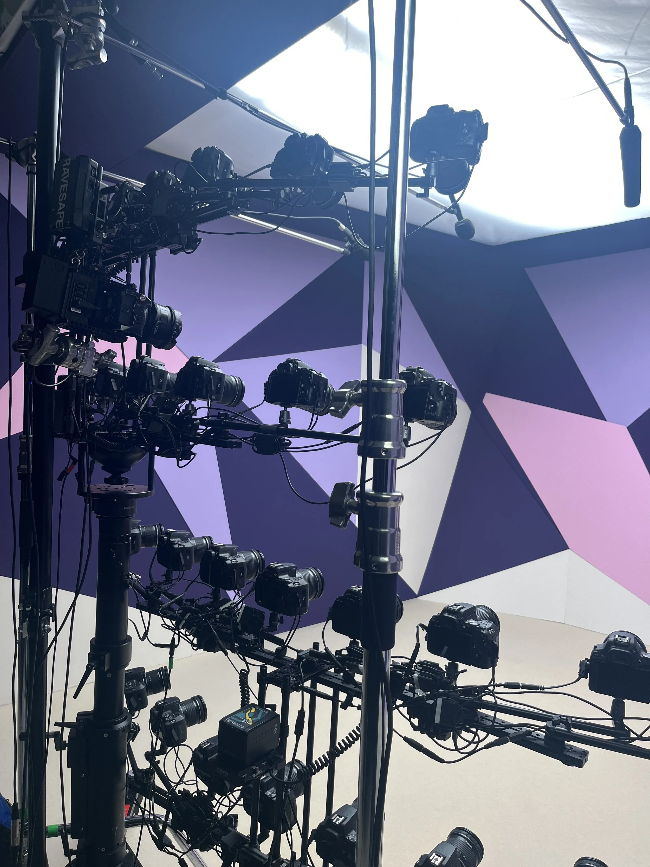 Multiple cameras mounted on rigs and stands inside a studio with a geometric purple and pink wall in the background.
