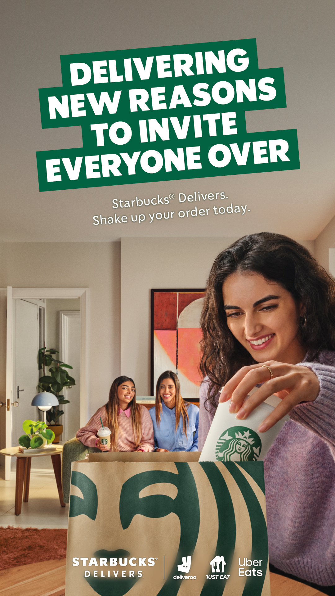 A woman in a pink sweater smiling and placing a Starbucks coffee cup into a paper bag with Starbucks and delivery service logos. In the background, two women sit on a couch, with one holding a Starbucks drink, in a cozy living room.