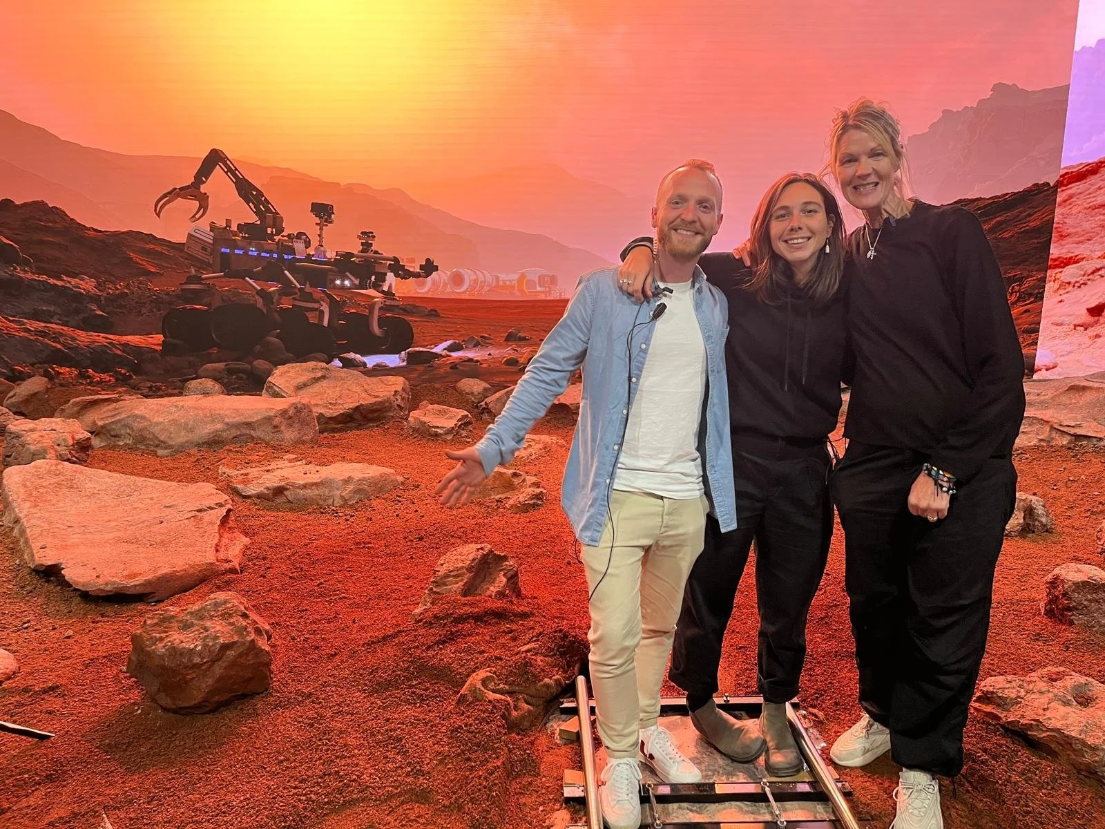 Three people, two women and one man, are standing on a set of tracks on a Mars-like landscape with reddish soil and rocks, smiling and posing for a photo. Behind them is a backdrop of a Martian terrain with a robotic rover and mountainous formations under a sunset sky.