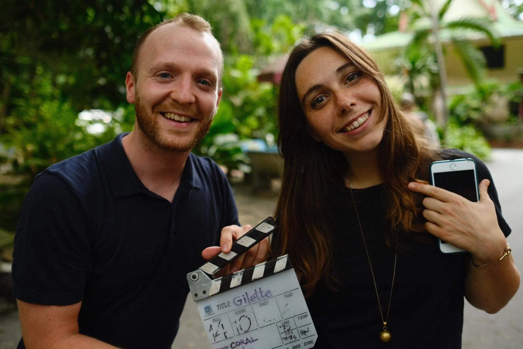 Two smiling people, a man and a woman, outdoors with greenery in the background. The man is holding a film clapperboard and the woman is holding a smartphone.