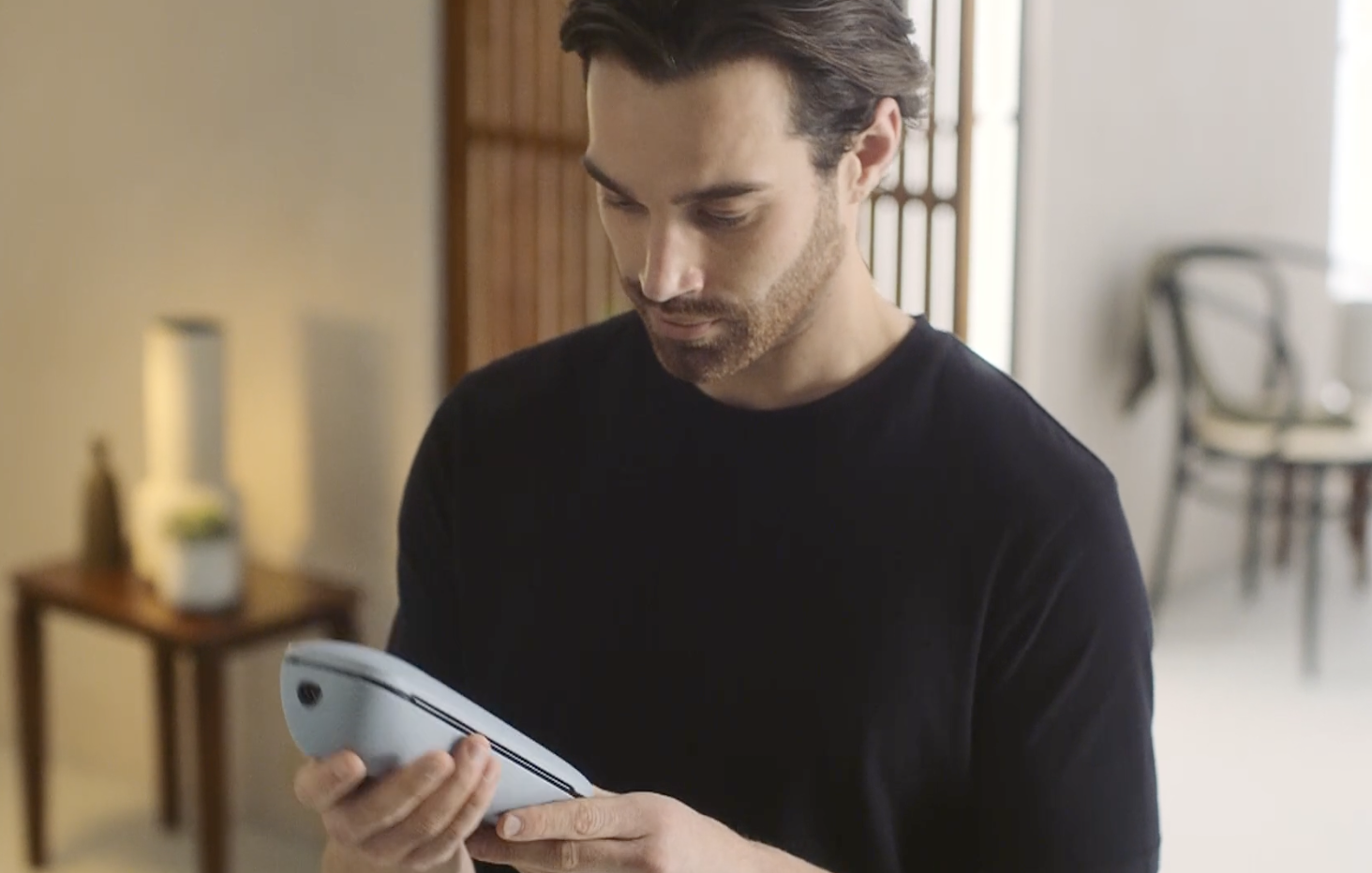 A man wearing a black shirt looking at a white electronic device in a room with natural light.