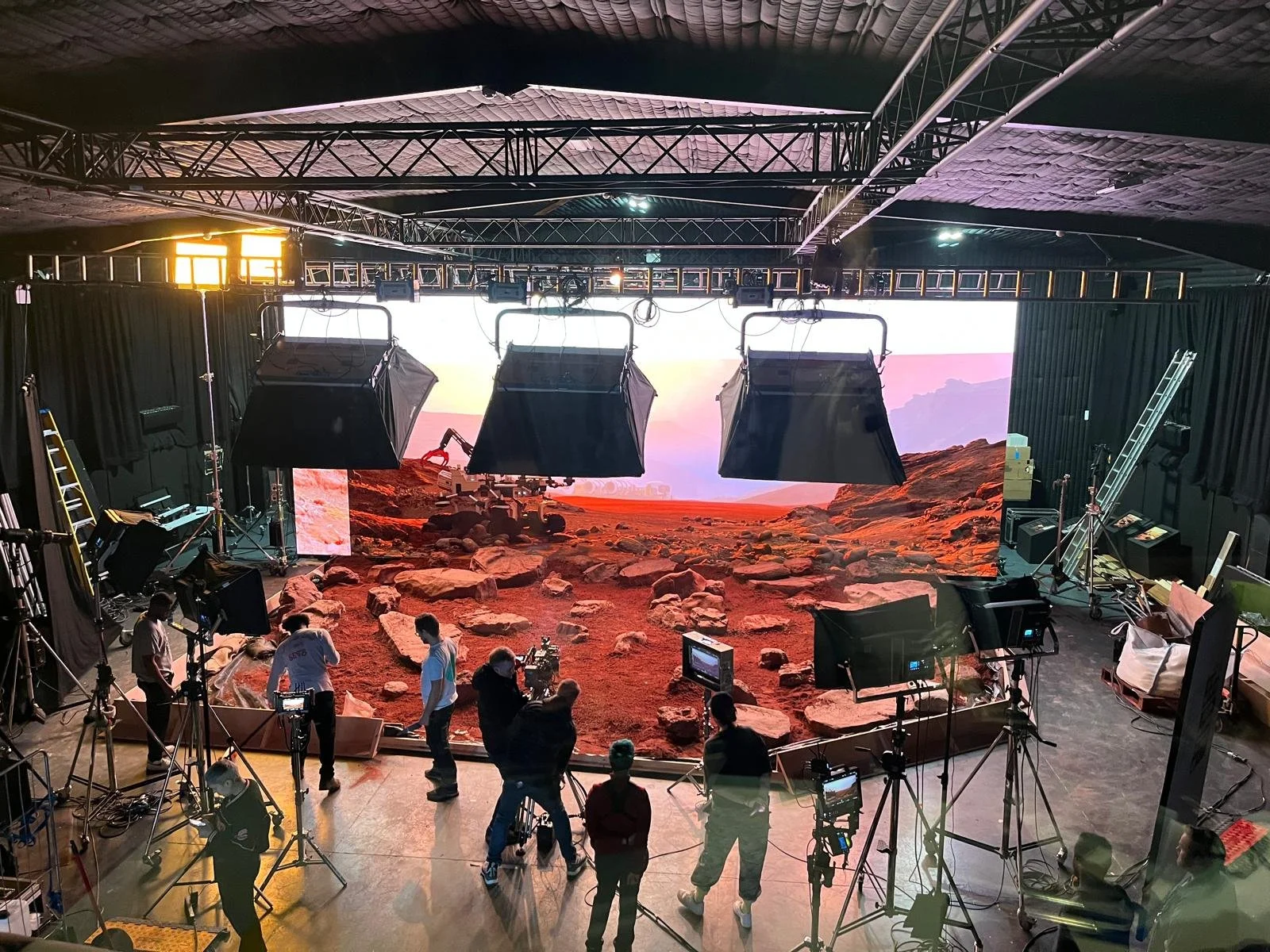 Film or television studio set with a Mars-like landscape backdrop, multiple production lights, cameras, crew members, and equipment.