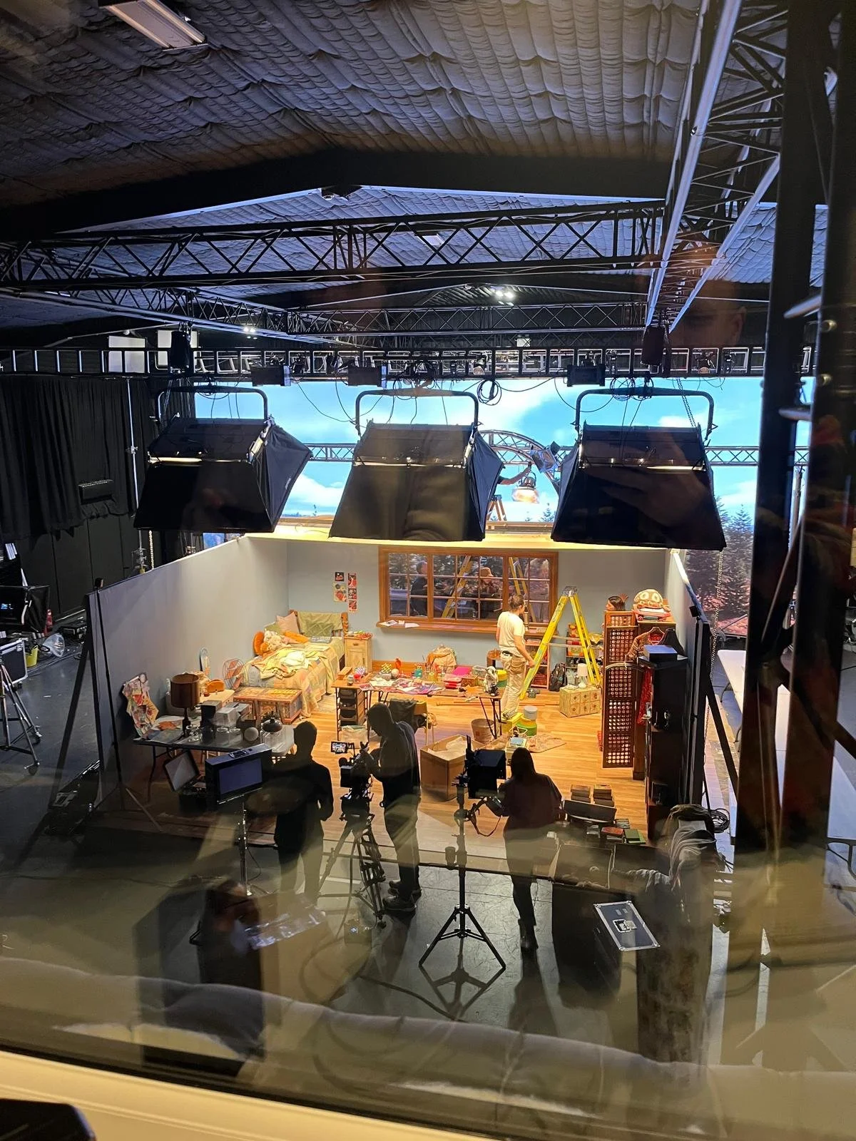 View of a filming set from behind a glass window, showing a scene with a cozy living room setup, including a sofa and chaotic toys, with crew members working and filming equipment around.
