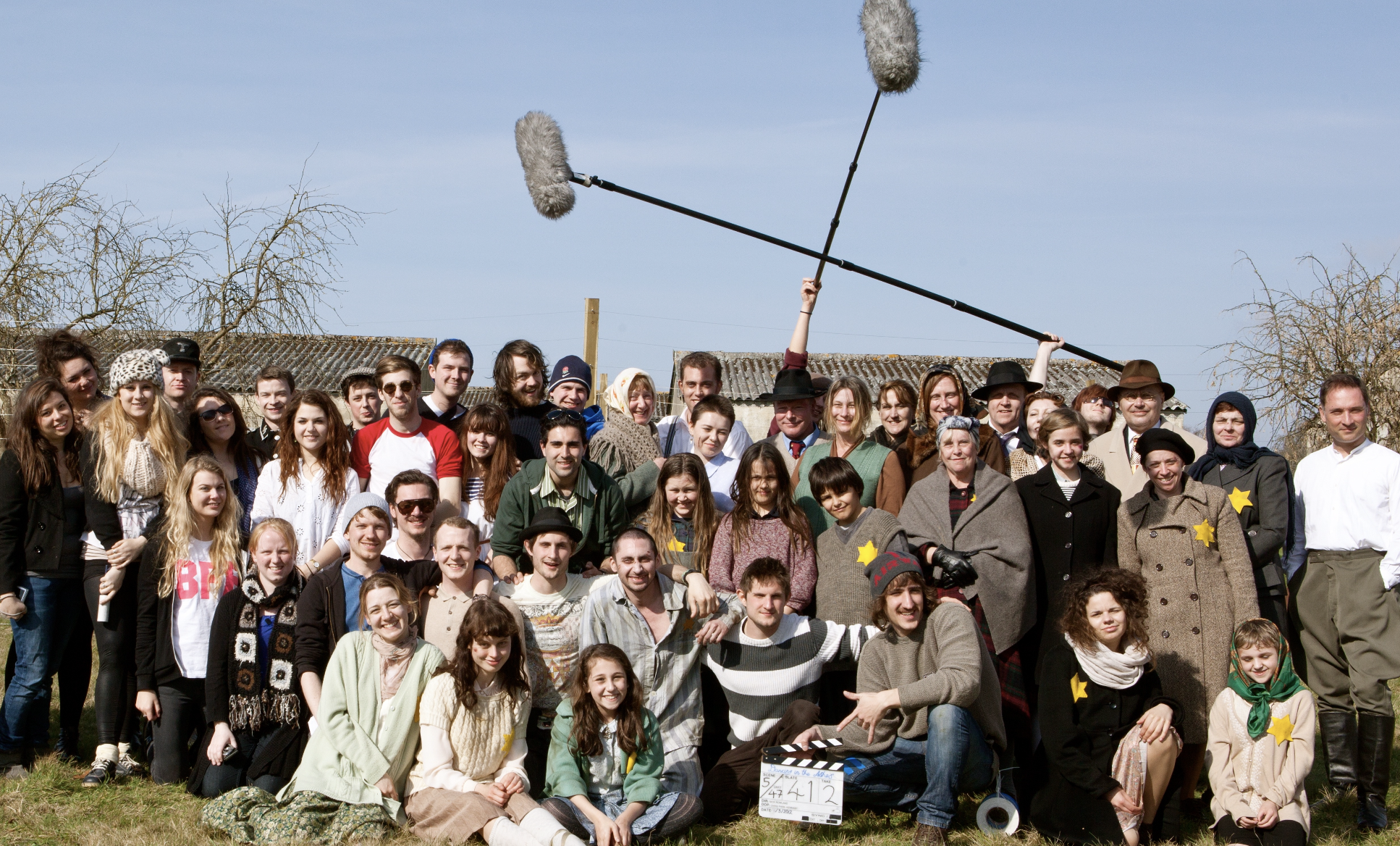 Group of people outdoors, some wearing vintage clothing, with a woman holding a long boom microphone above their heads, on a sunny day with a clear sky.