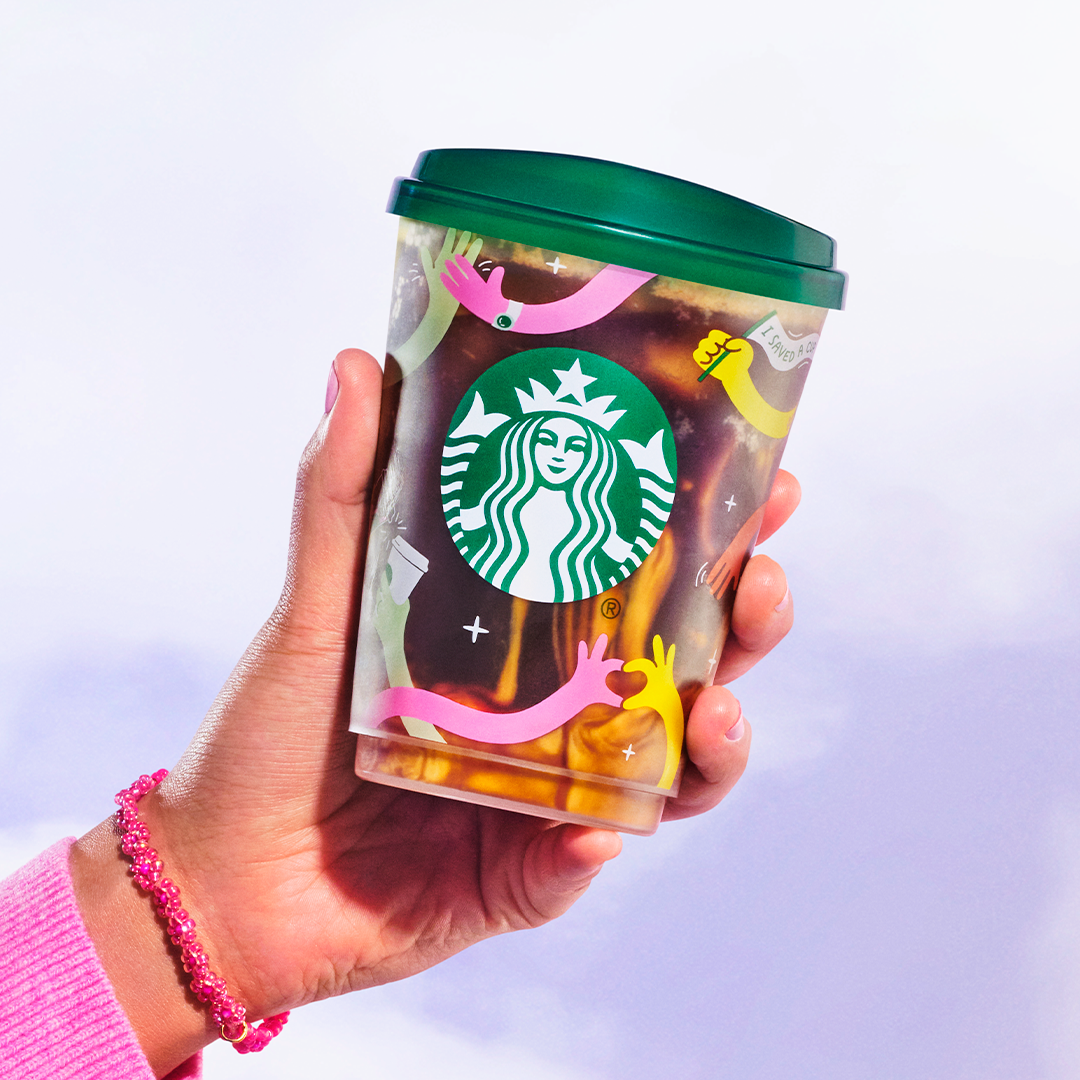 A hand holding a Starbucks coffee cup with colorful sticker illustrations of hands in various colors and a small message sticker that says 'I saved a' with the rest of the message obscured. The cup has a green lid and the background is plain and ligh