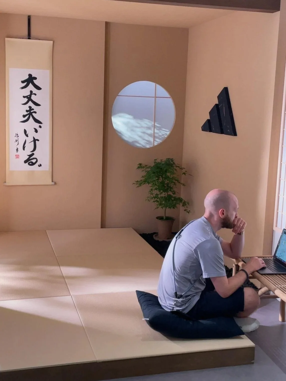 A man sitting on a cushion on a wooden floor, working on a laptop in a room with Japanese-style decor, including a hanging scroll with Japanese characters, a round window showing clouds, and a potted plant.