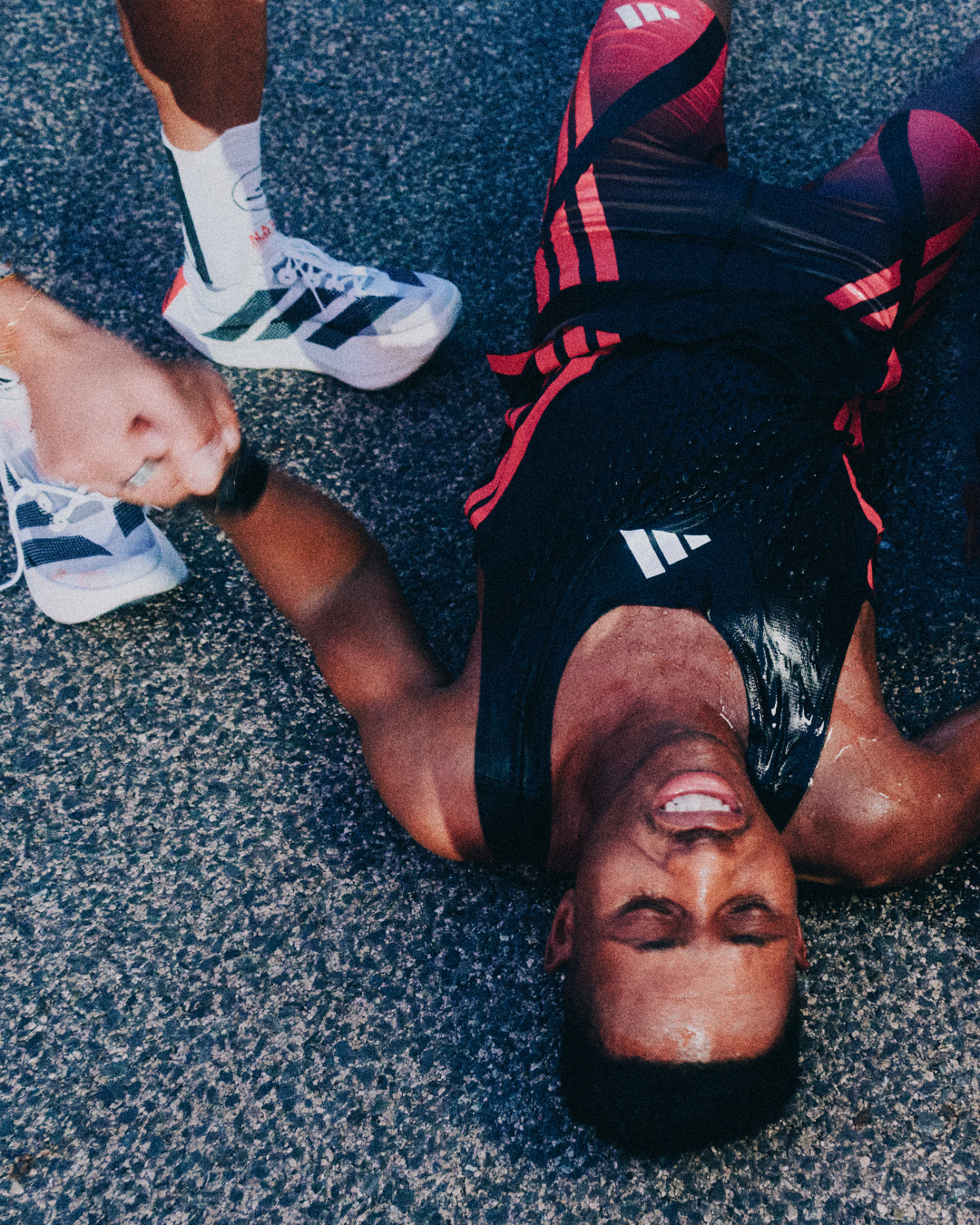 A man is lying on the ground after a workout or run, smiling with his eyes closed, wearing black and red athletic clothes.