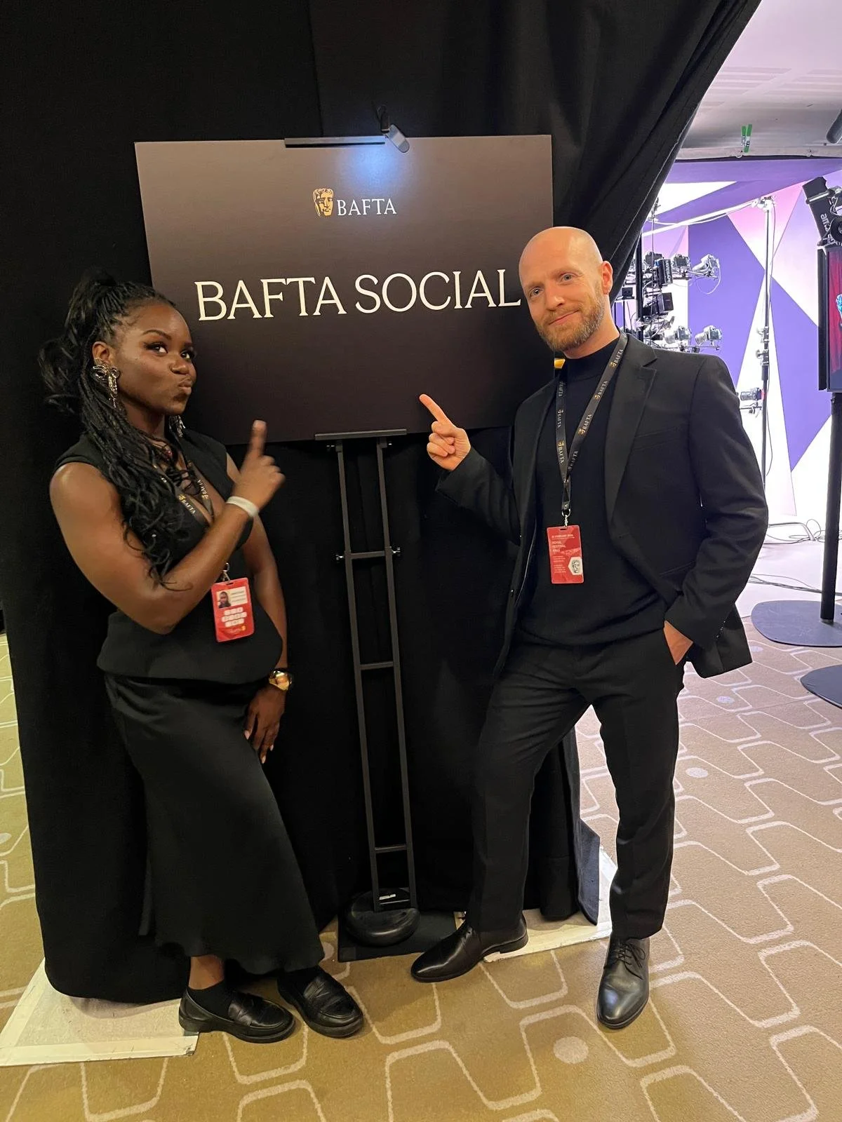 A woman and man standing beside a sign that reads "BAFTA SOCIAL" at an event, both pointing at the sign. The woman has long curly hair, dark clothing, and is wearing an event badge. The man is bald, with a beard, black suit, and event badge. There is a dark background with professional lighting and equipment.