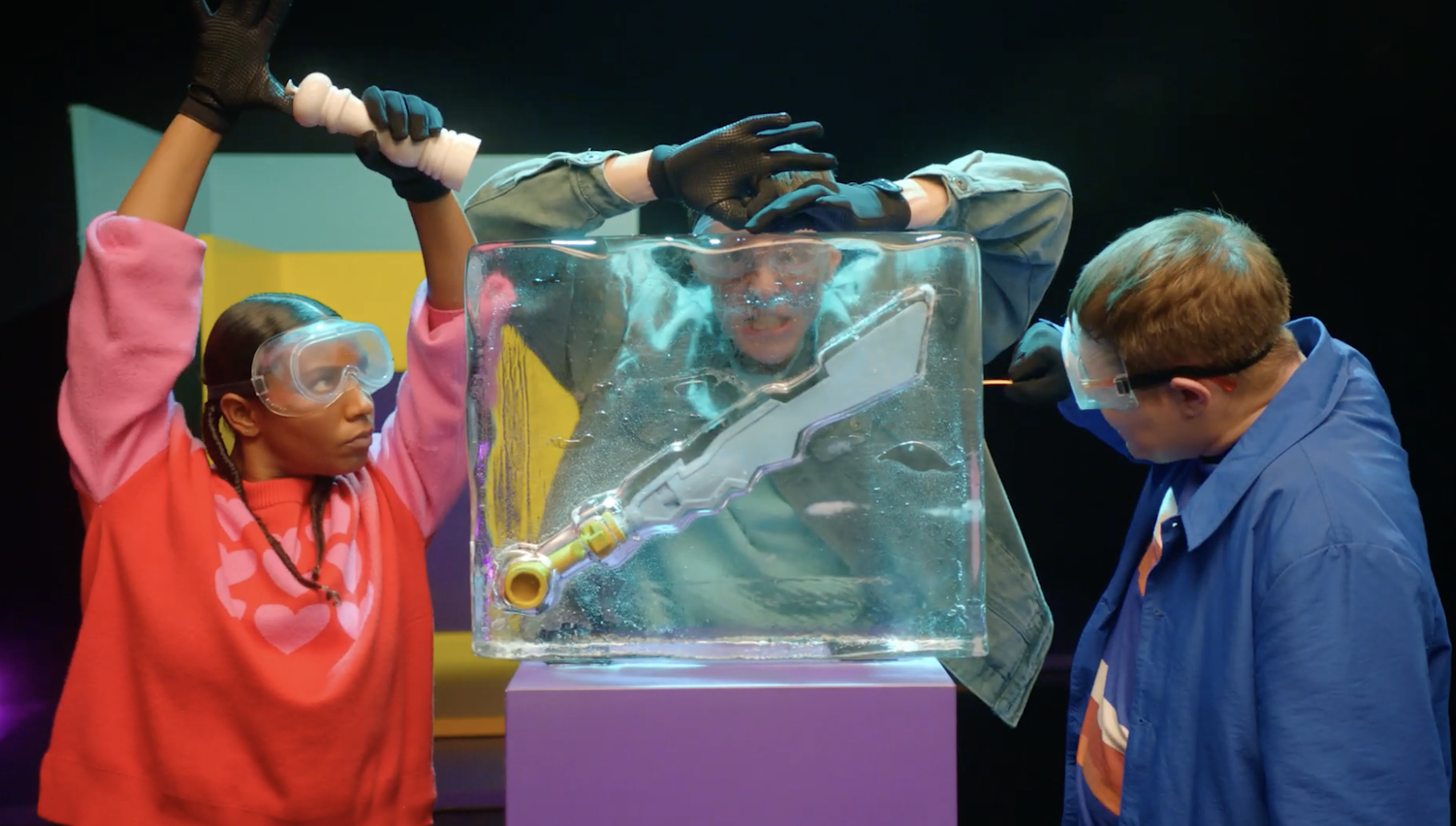 Two children, a girl and a boy, are wearing protective goggles and gloves, engaging in an experiment with a large, clear ice block containing a model of a sword. They are in a science exhibit or laboratory setting with colorful background panels.