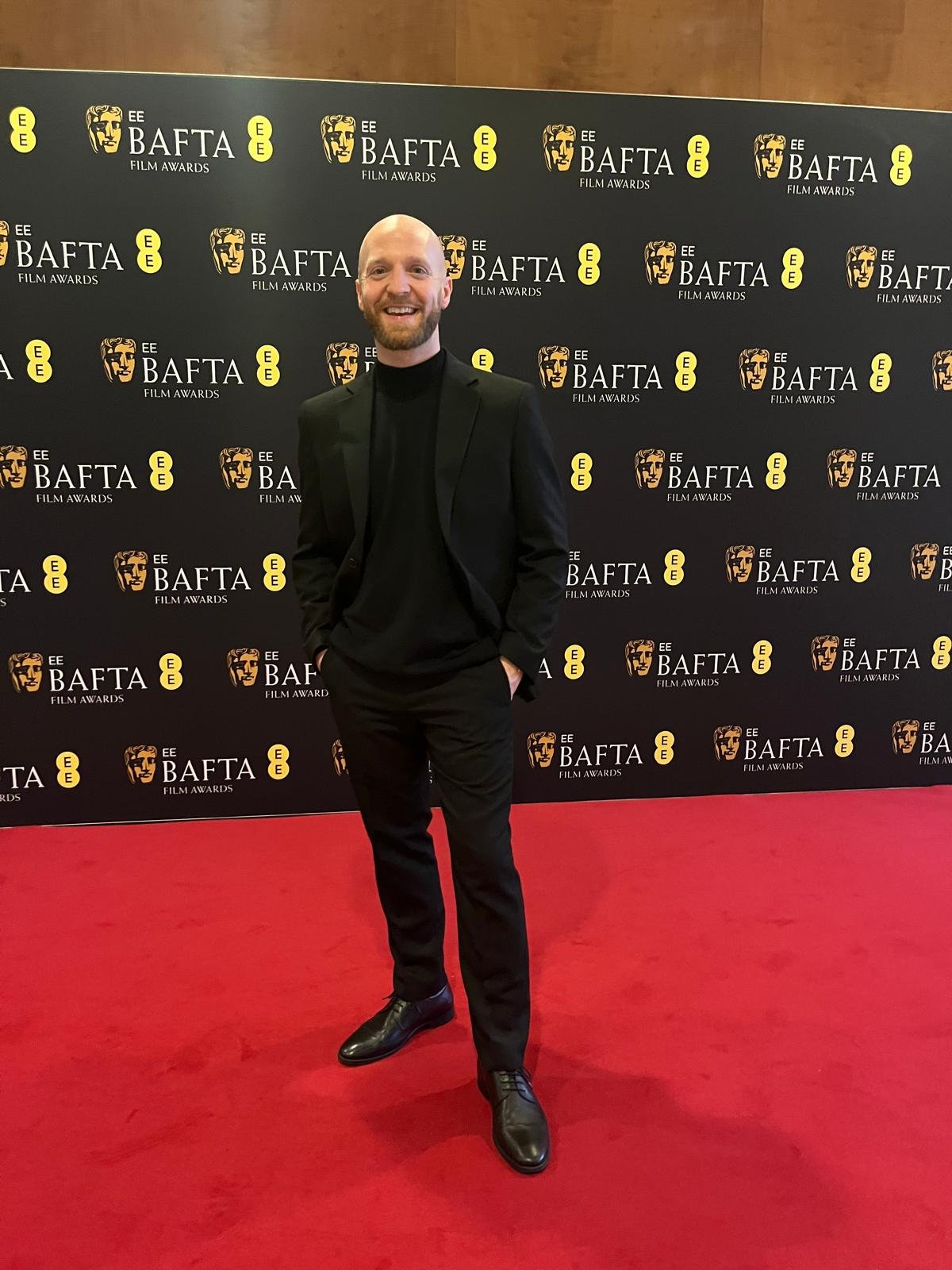 A man in a black suit standing on a red carpet at the BAFTA Film Awards, with a BAFTA backdrop behind him.