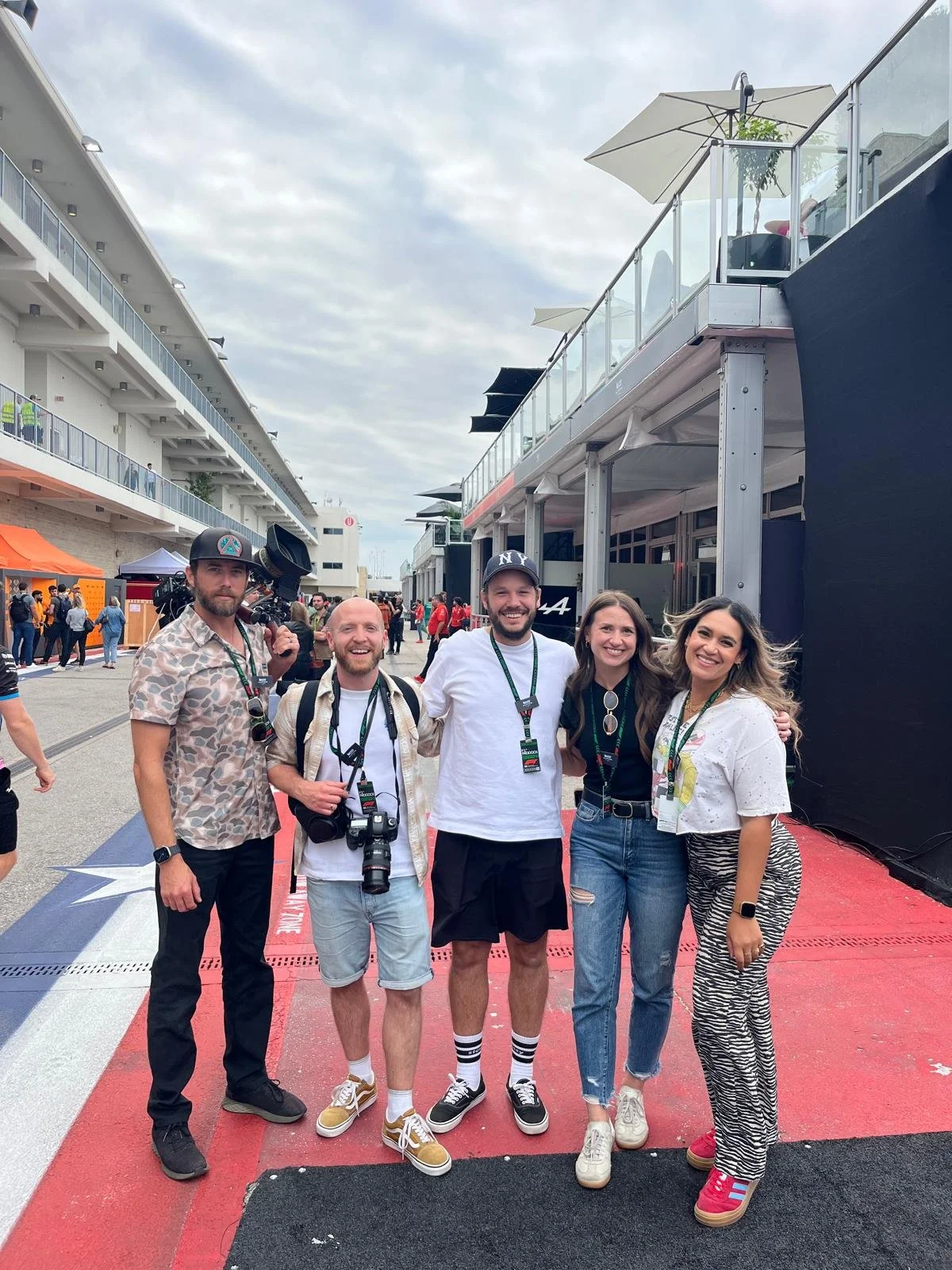 A group of five people smiling and posing for a photo at a racing event, with race track and spectators in the background.