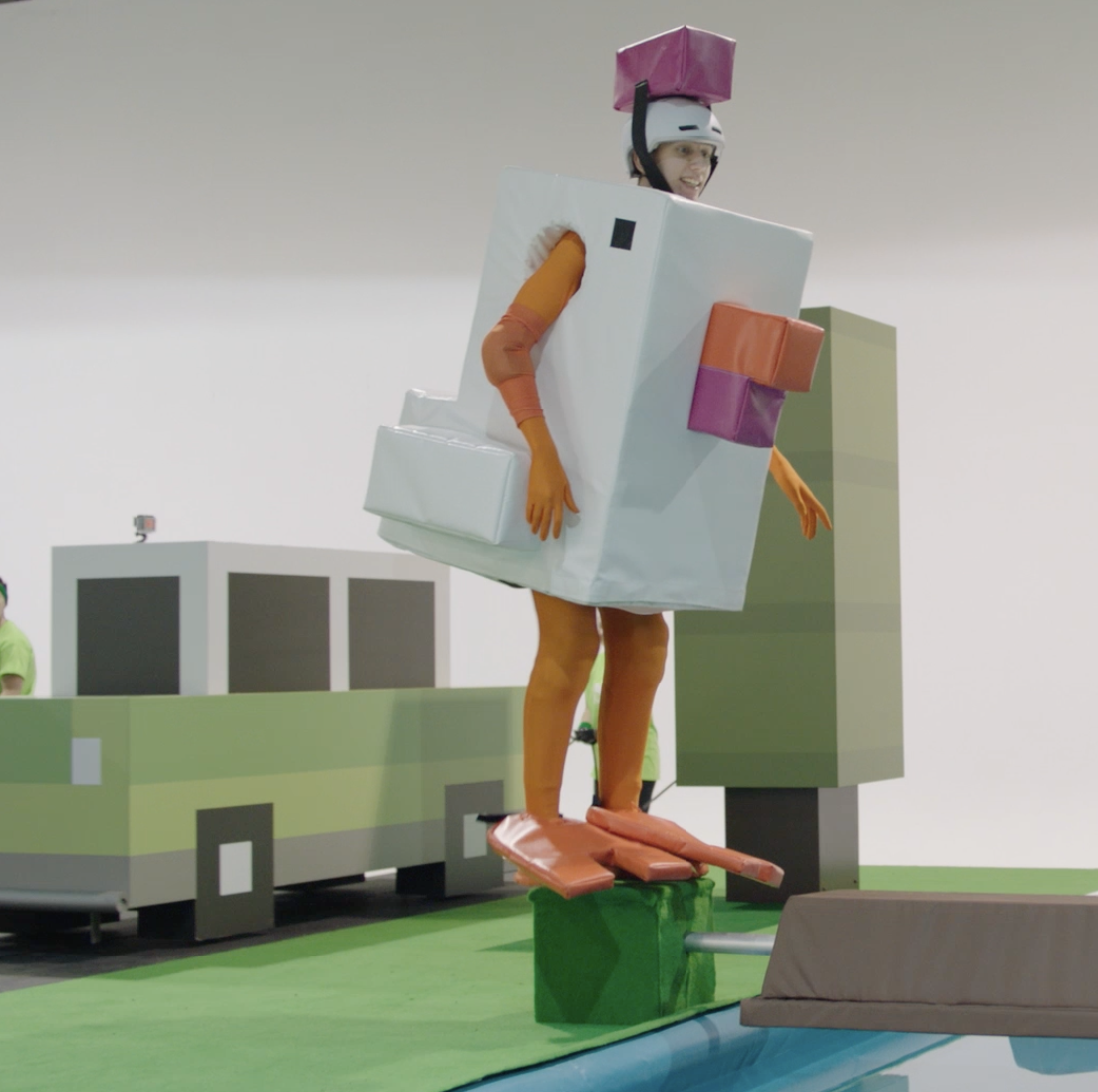 Person dressed as a large gift box with colorful bows, wearing a helmet, standing on a green platform.