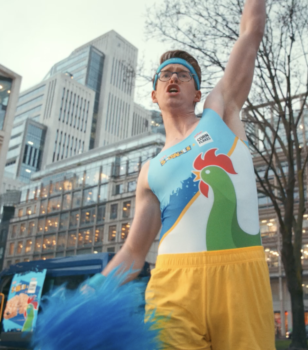 A man wearing glasses and sportswear with a rooster graphic, yellow shorts, and a blue headband, raising his arm outdoors in an urban area with tall buildings and leafless trees.