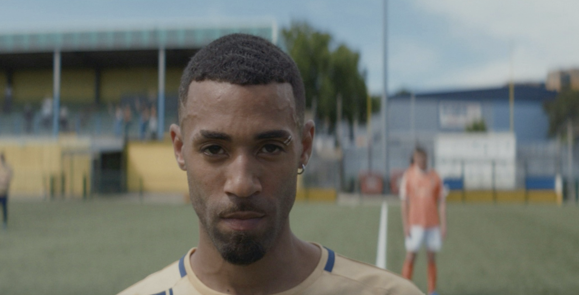 Close-up of a young man with a serious expression standing on a soccer field, with a person in orange sportswear in the background.