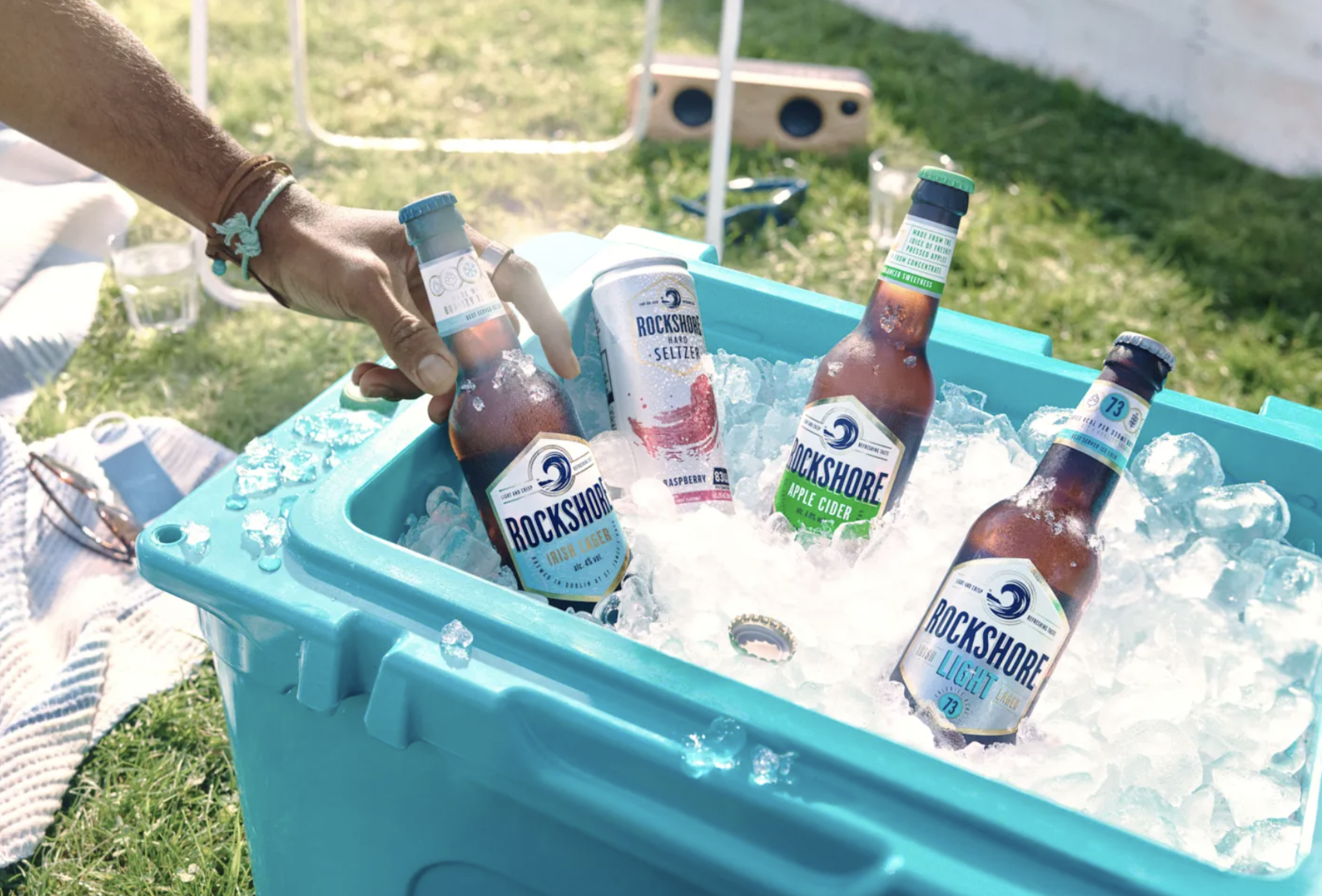 A person is placing a bottle of Rockshore Irish Lager into a blue cooler filled with ice, containing four other bottles of Rockshore beverages, on a grassy outdoor setting.