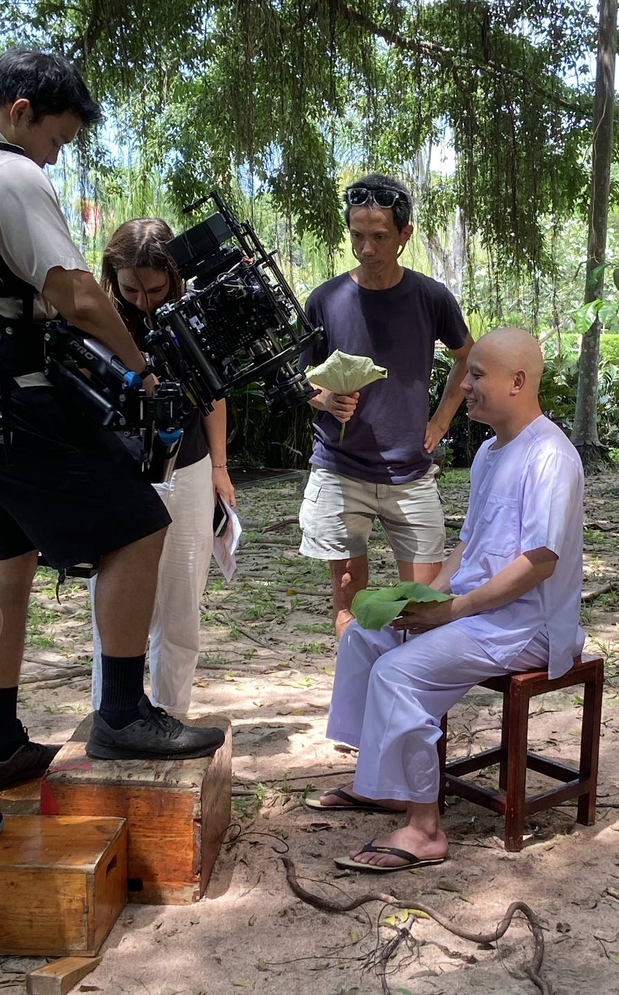 Filmmaking crew filming a scene in a jungle, with a man in white sitting on a small stool holding a green leaf, and others standing around with equipment.