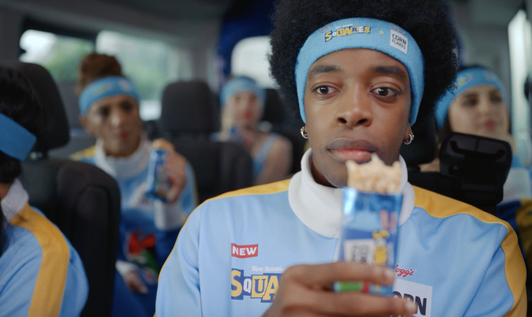 A person with curly hair wearing a blue headband and a matching light blue and yellow sports uniform is sitting on a bus, holding a snack bar. Several other individuals in similar uniforms are in the background, also wearing blue headbands and eating snacks.