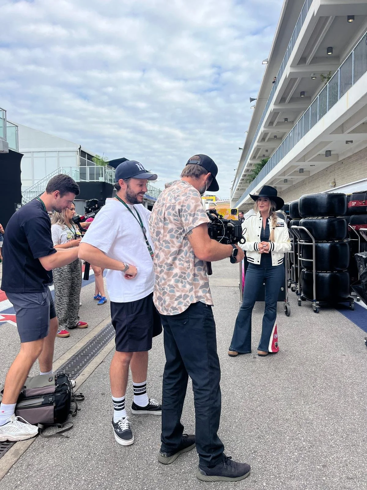 Group of people filming a woman in a white jacket and wide-brimmed hat at a racing event, with tire stacked on the side and a multi-story building in the background.