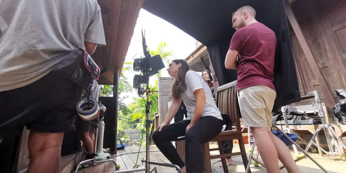 Woman sitting on a chair during a film shoot, surrounded by crew members operating camera equipment, outdoors in a wooden structure.