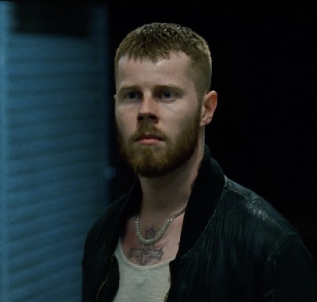 A man with red hair and beard looking sideways, standing next to a blue wall