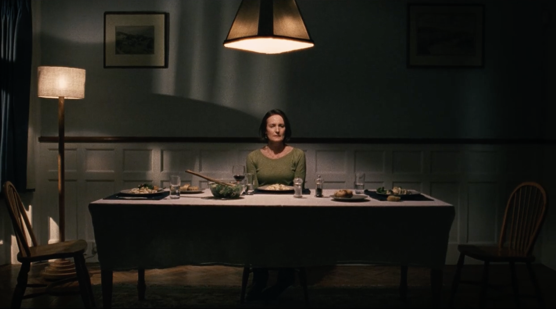 A woman with dark hair sits alone at a dinner table in a dimly lit room, with a lampshade overhead, surrounded by plates of food, glasses, and cutlery.