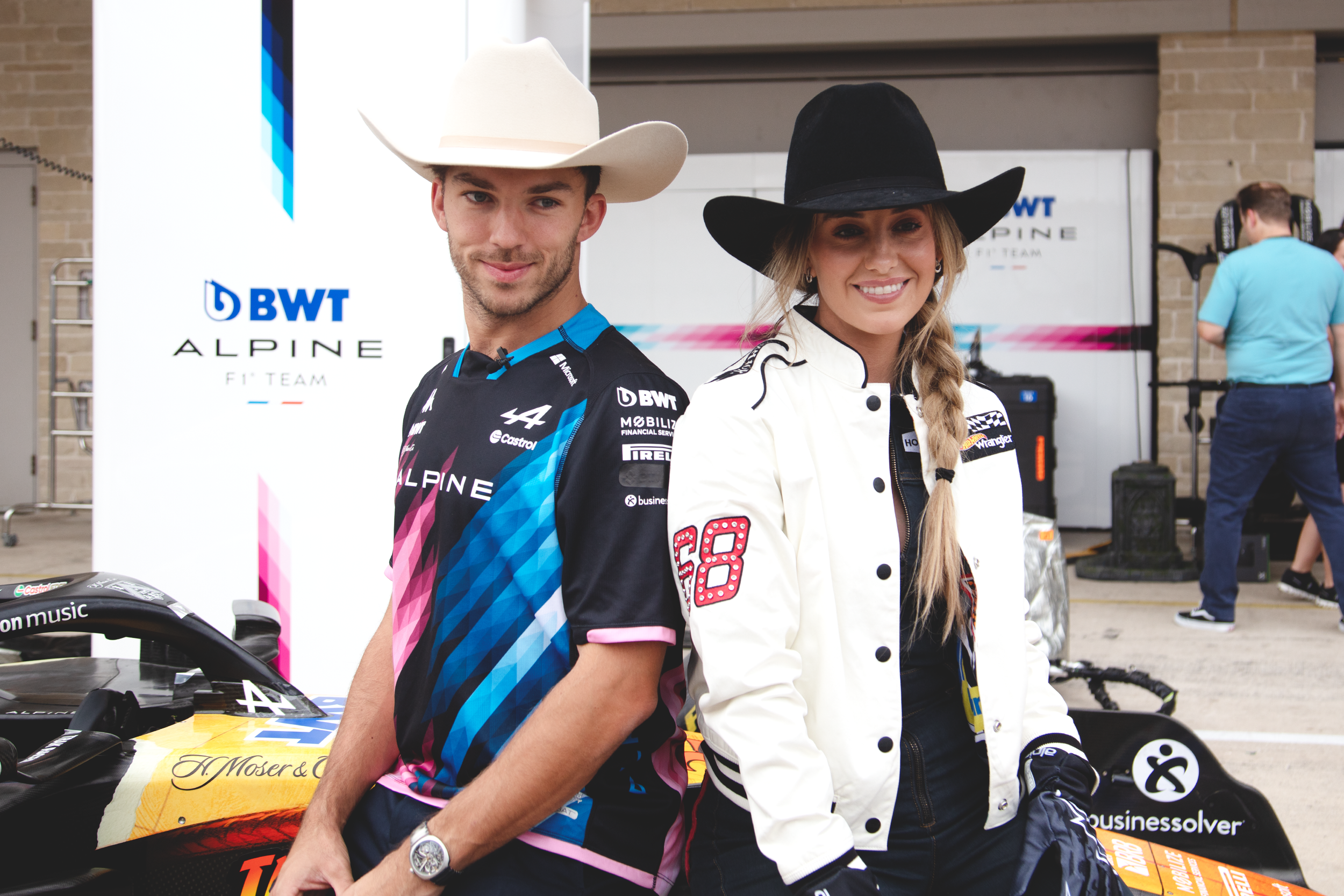 Two race car drivers, a man and a woman, standing next to a race car in a garage area. The man is wearing a black and blue Alpine race suit and a white cowboy hat, while the woman is wearing a white and black racing jacket and a black cowboy hat, both smiling.