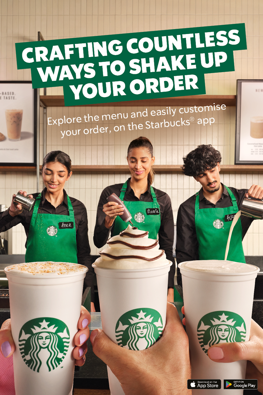 Three Starbucks employees behind the counter showing three cups of drinks with whipped cream and chocolate drizzle, with hands holding similar drinks in the foreground, and a sign about customizing orders with the Starbucks app.